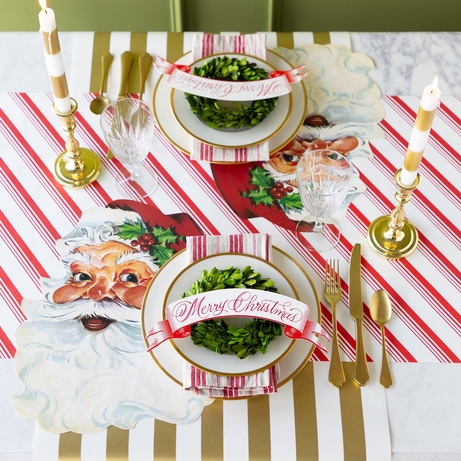Christmas-themed table setting with Die-cut Santa Claus Placemats, Merry Christmas Red Banner Table Accents, candles, topiary wreaths and gold flatware on the Peppermint Stripe and Gold Classic Stripe Runners. 