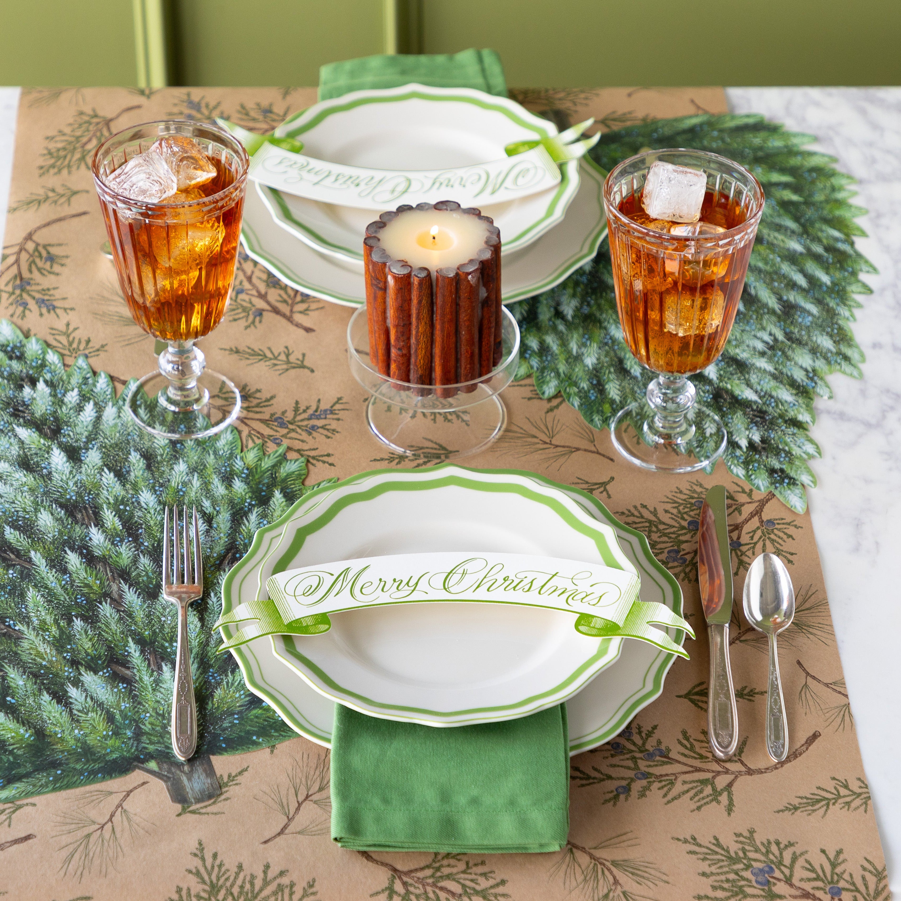 Winter table setting with green and white scalloped plates, Moss Merry Christmas Banner Table Accent and flatware on the Die-cut Evergreen Placemat and Juniper Sprigs Runner.