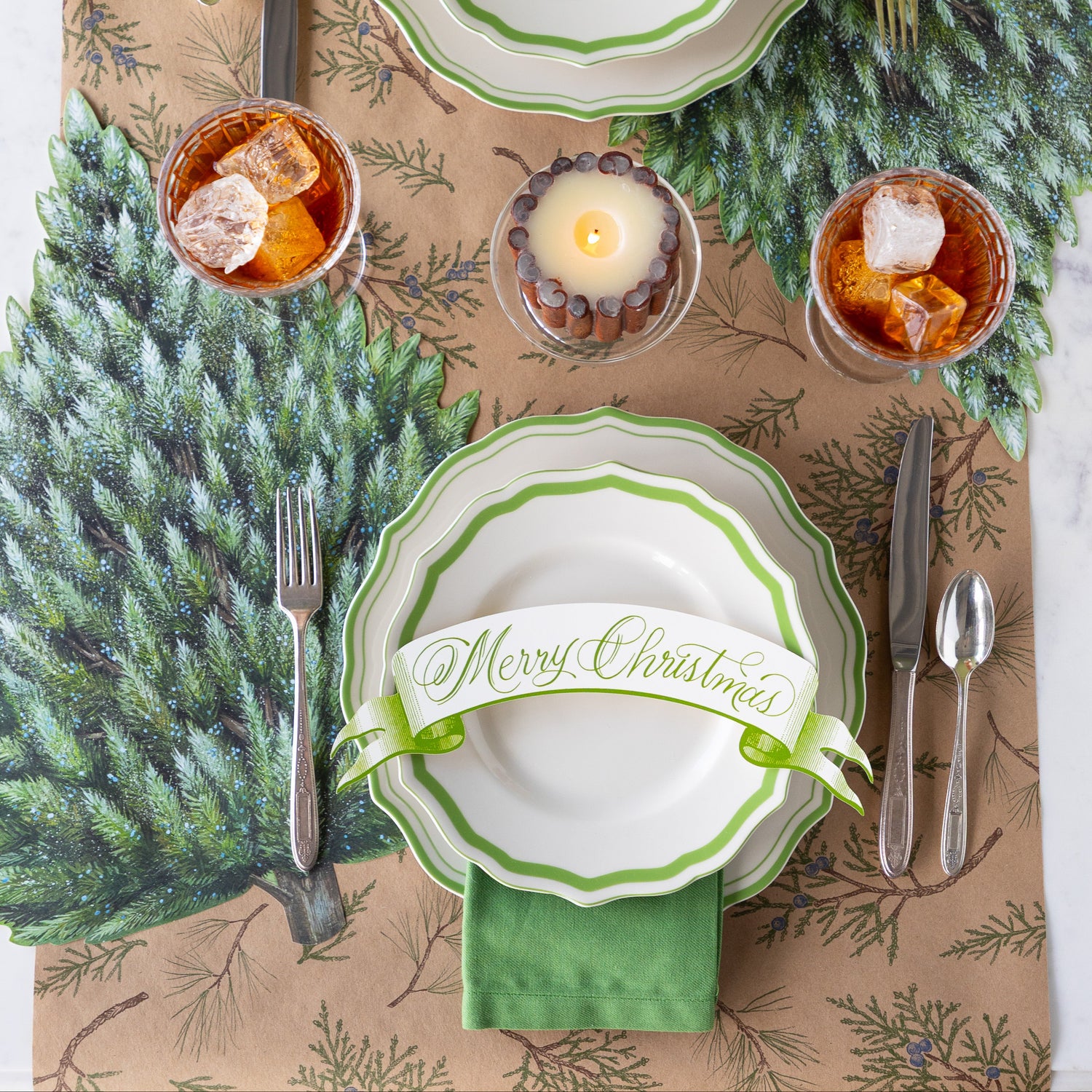 Winter table setting with green and white scalloped plates, Moss Merry Christmas Banner Table Accent and flatware on the Die-cut Evergreen Placemat and Juniper Sprigs Runner.