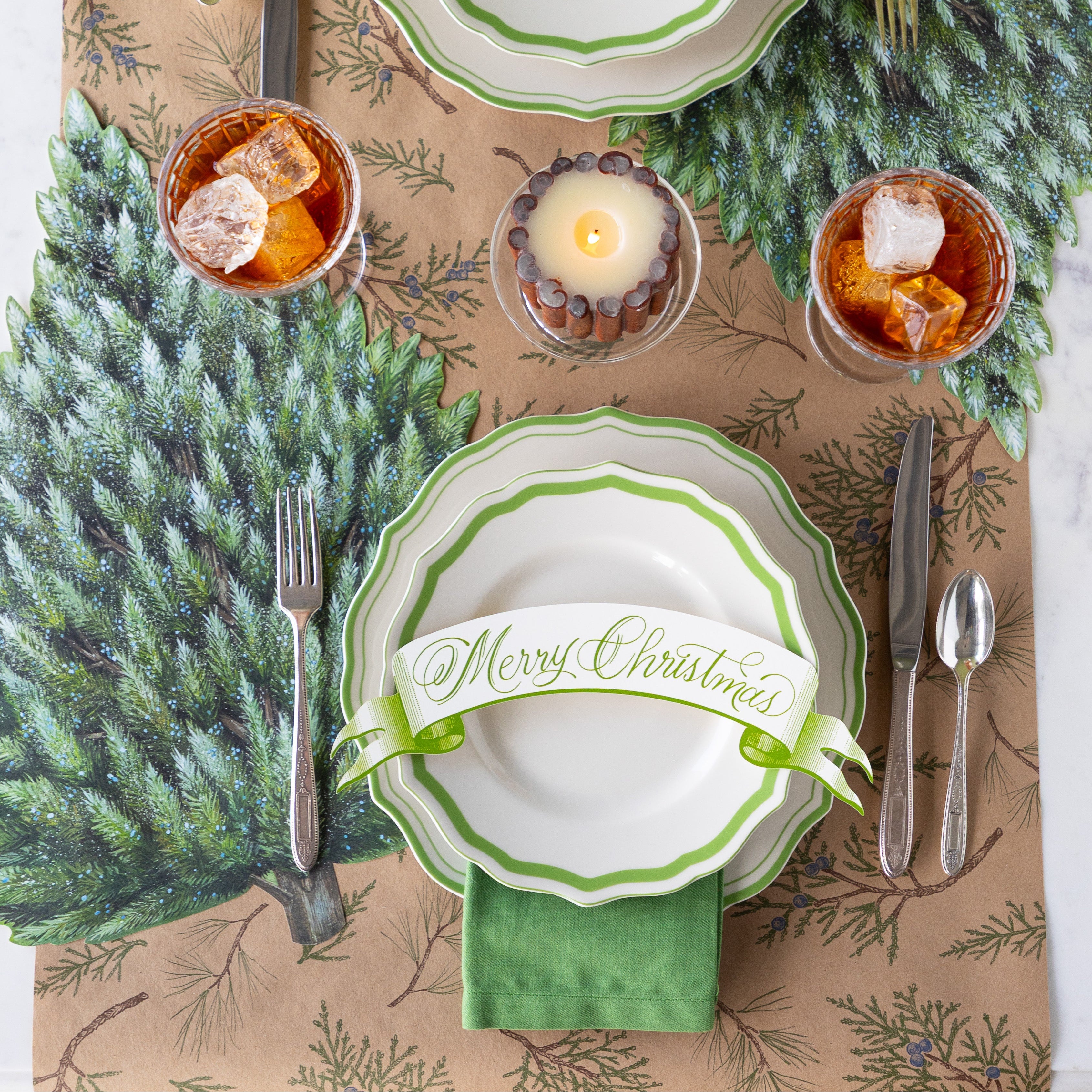 Winter table setting with green and white scalloped plates, Moss Merry Christmas Banner Table Accent and flatware on the Die-cut Evergreen Placemat and Juniper Sprigs Runner.