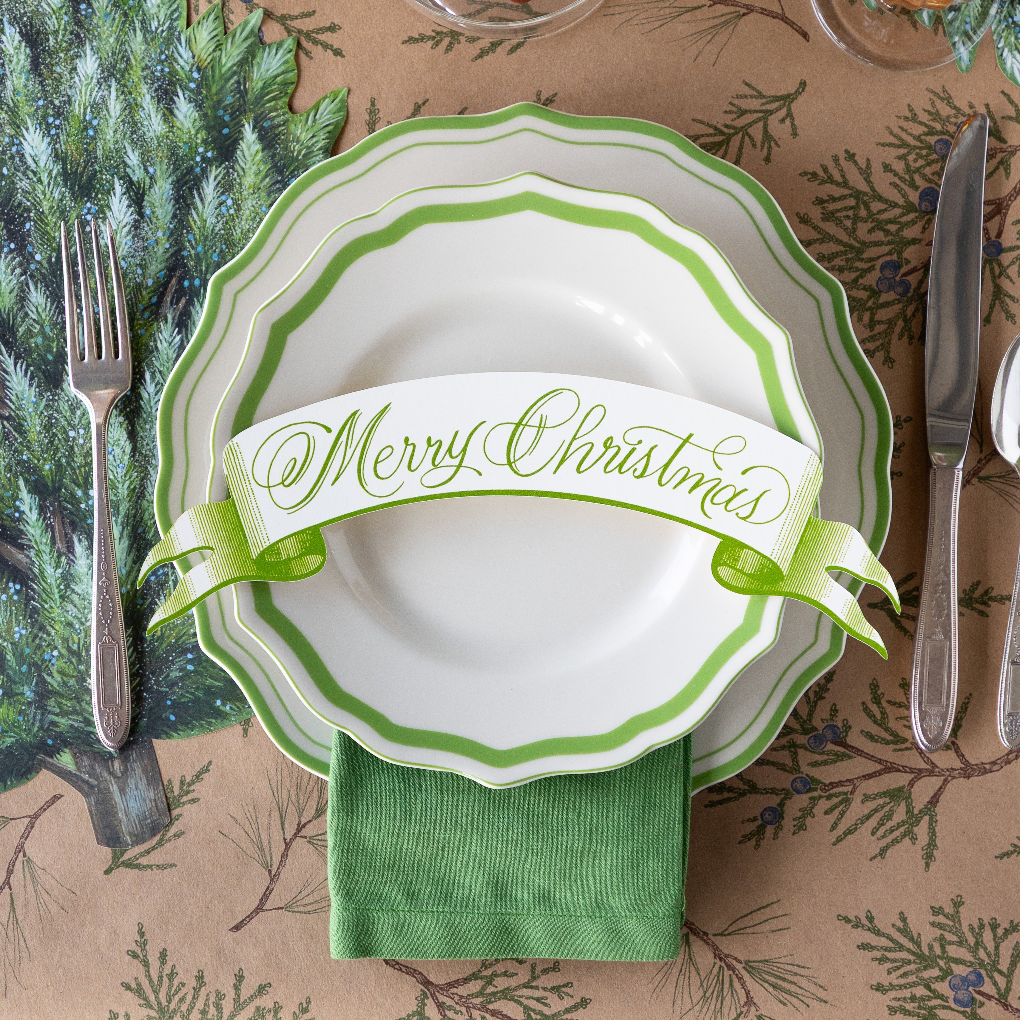 Winter table setting with green and white scalloped plates, Moss Merry Christmas Banner Table Accent and flatware on the Die-cut Evergreen Placemat and Juniper Sprigs Runner.