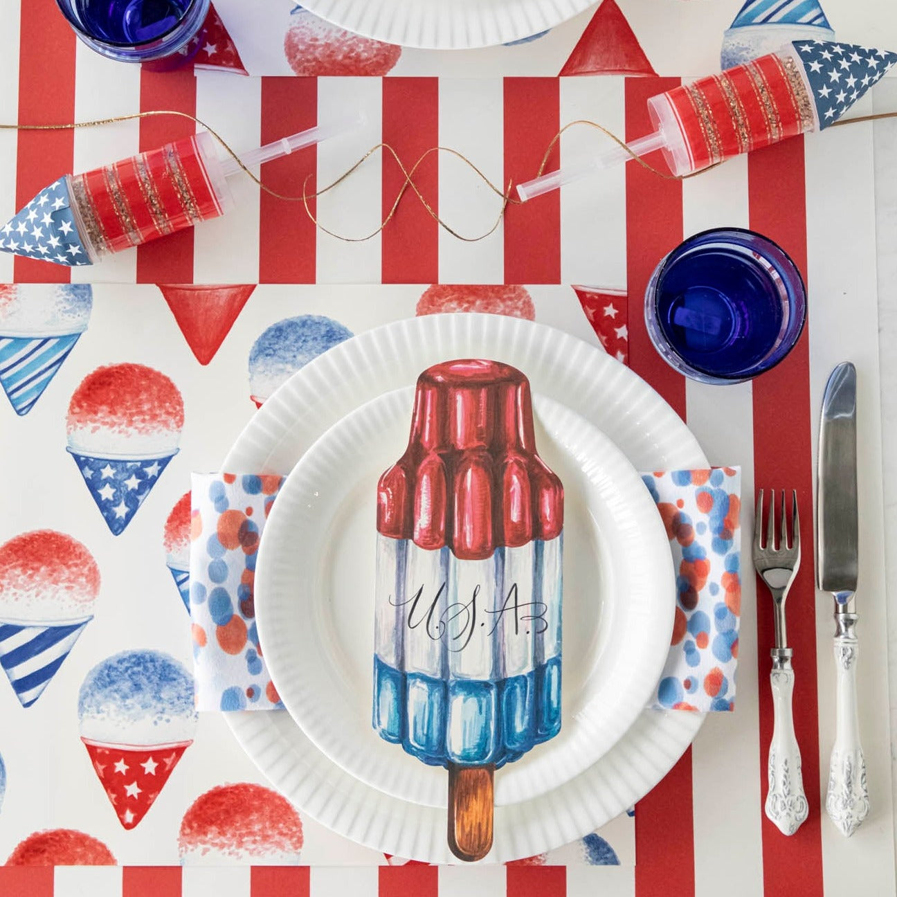 Celebrate the 4th of July with a patriotic table setting featuring a Snow Cone Placemat adorned with red, white, and blue toppings. Complete the summer-themed ambiance with a USA flag.