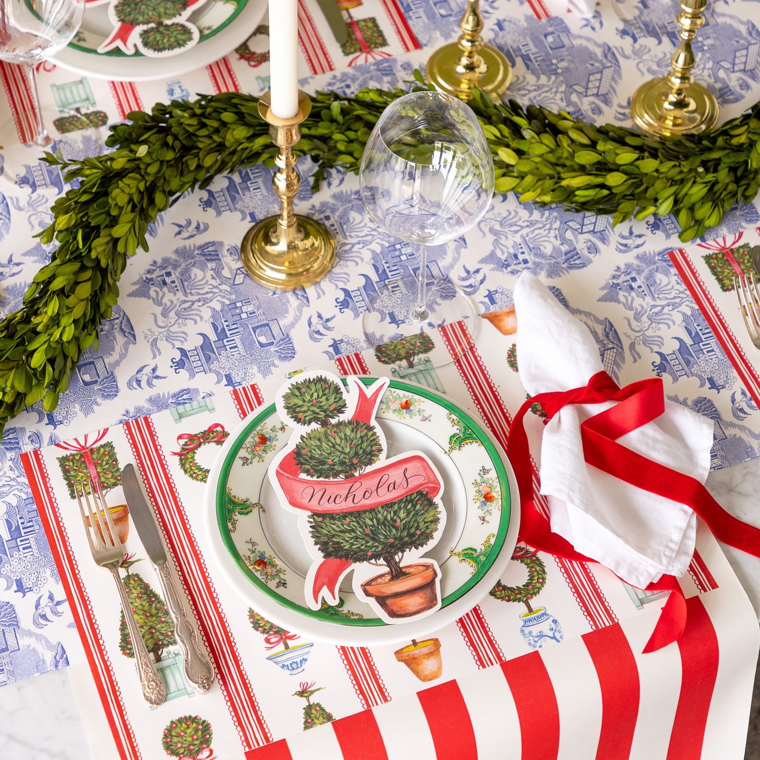 An elegant table setting with a topiary garland featuring the Holiday Topiary Placemat, a red ribbon tied around a napkin and Red Classic Stripe Runner running vertically and Blue Regal Peacock Runner running horizontally underneath.