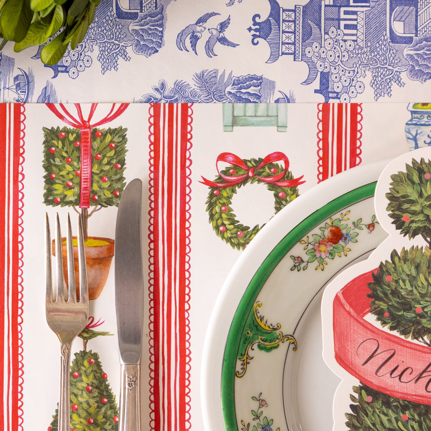 A place setting featuring the Holiday Topiary Placemat, a fork and knife, elegant dinnerware and Blue Regal Peacock Runner underneath.