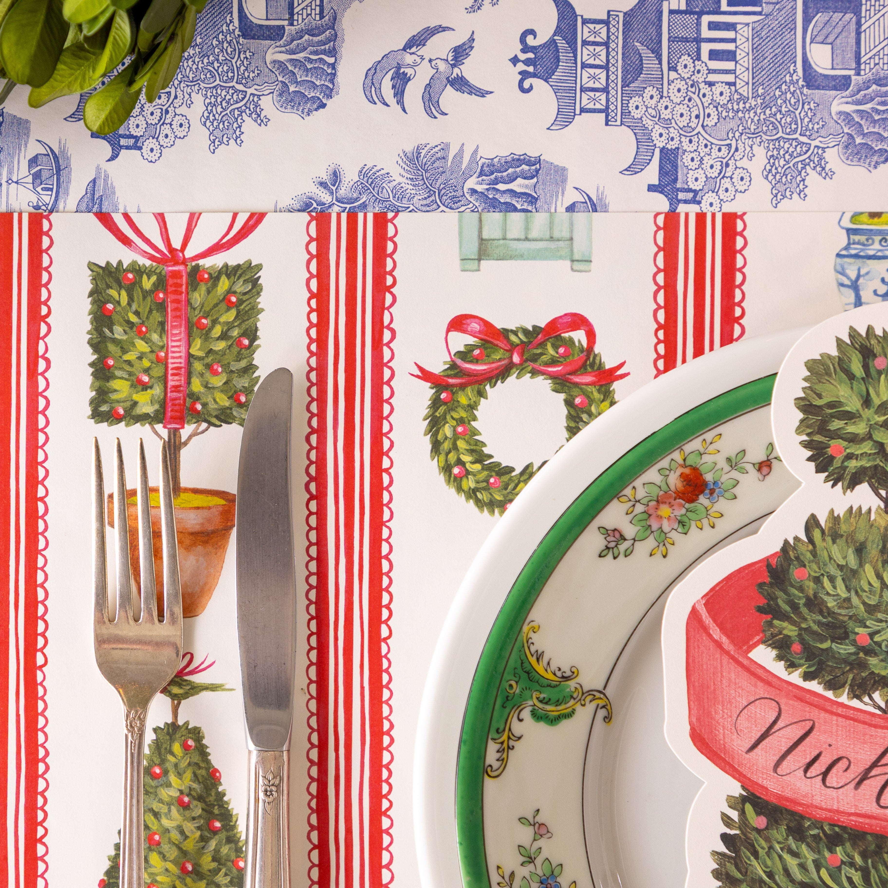 A place setting featuring the Holiday Topiary Placemat, a fork and knife, elegant dinnerware and Blue Regal Peacock Runner underneath.