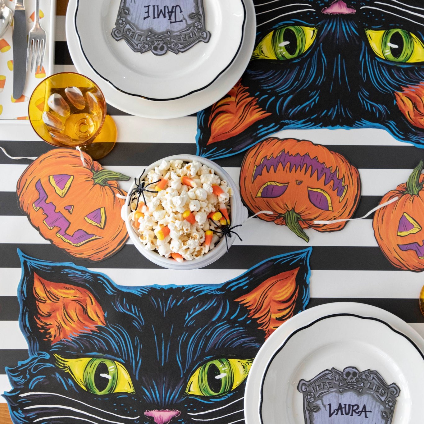 Overhead view of a spooky tablescape featuring the Die-cut Black Cat Placemat, pumpkin bunting, a bowl of popcorn and candy corn with fake spiders on top and a Tombstone Place Card.