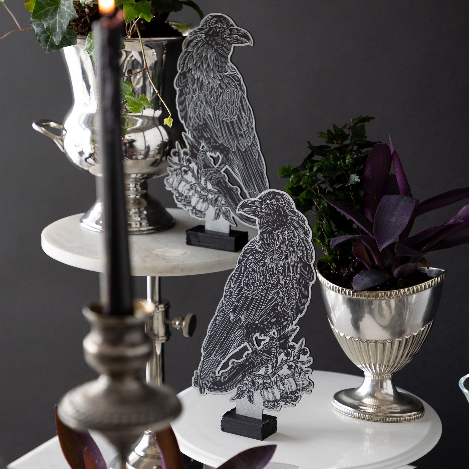 Two Raven Table Ornaments on cake stands of different heights, next to dark foliage.