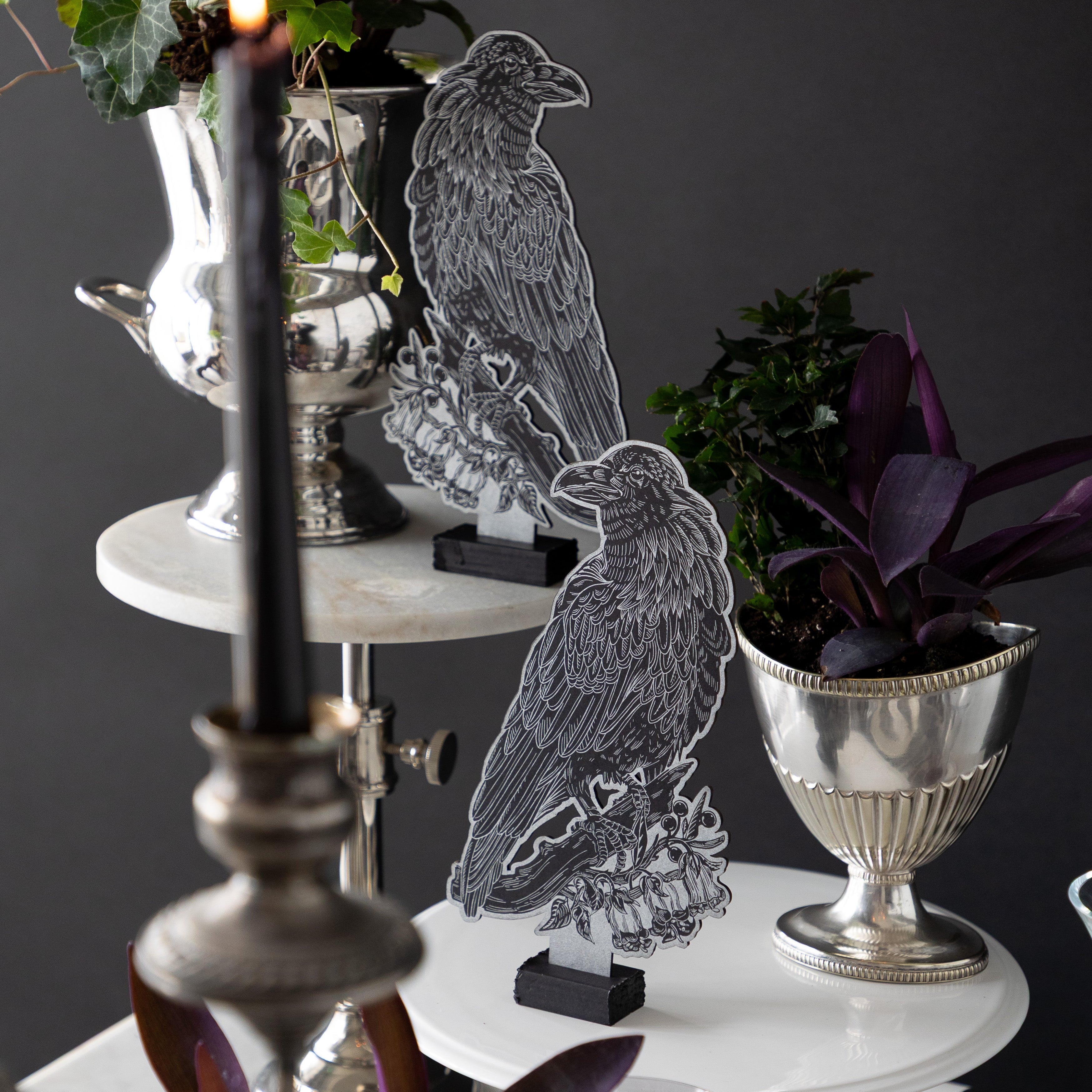 Two Raven Table Ornaments on cake stands of different heights, next to dark foliage.