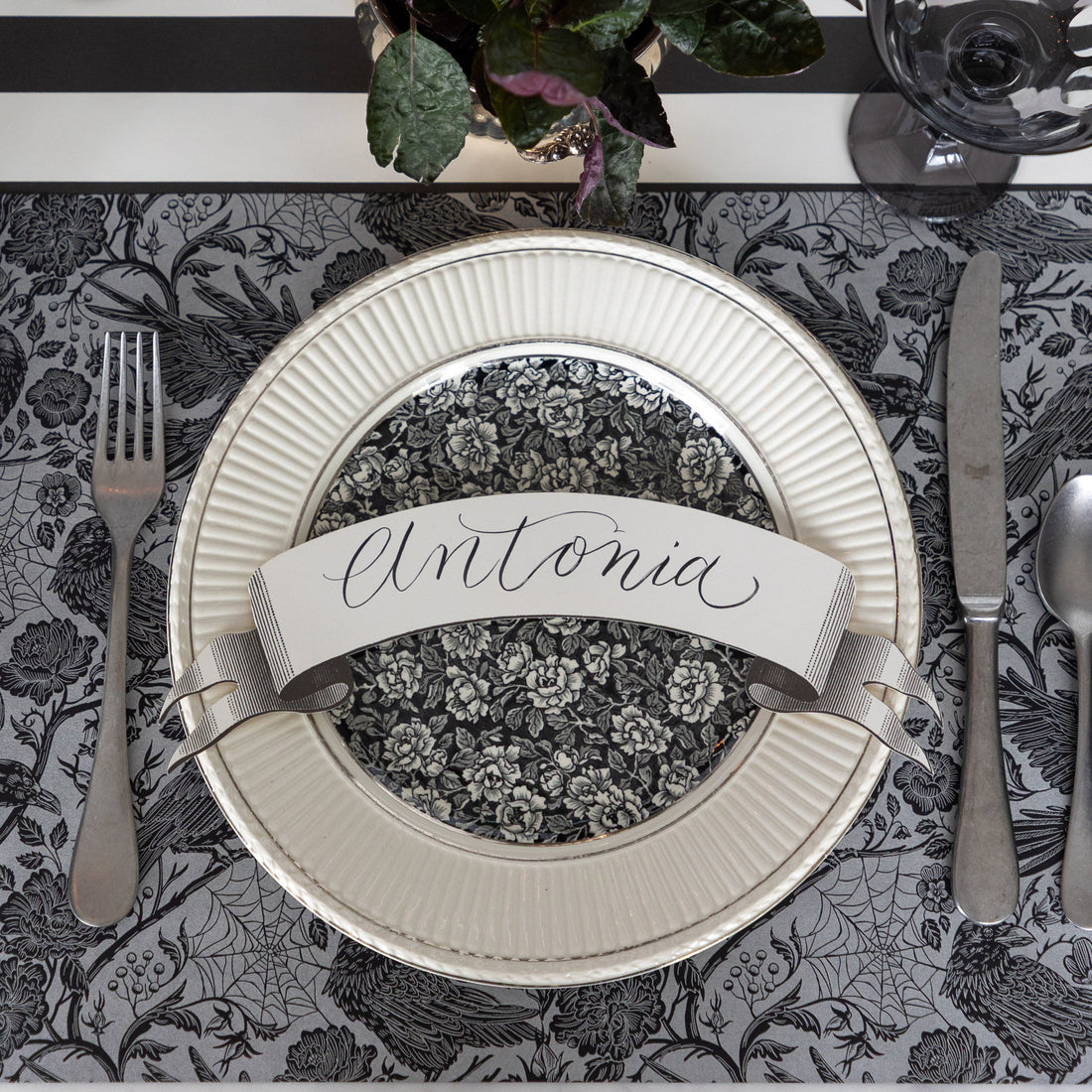 A spooky place setting featuring the Raven Toile Placemat with silver flatware, a dinner and salad plate with the Classic Black Banner Table Accent with "Antonia" written on it on top and Black Classic Stripe Runner underneath.
