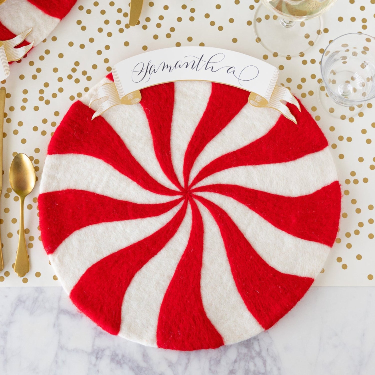 The Red Felt Candy Stripe Placemat with the Classic Gold Banner Table Accent reading &quot;Samantha&quot; atop, gold flatware beside it, an empty glass, and Gold Confetti Runner underneath.