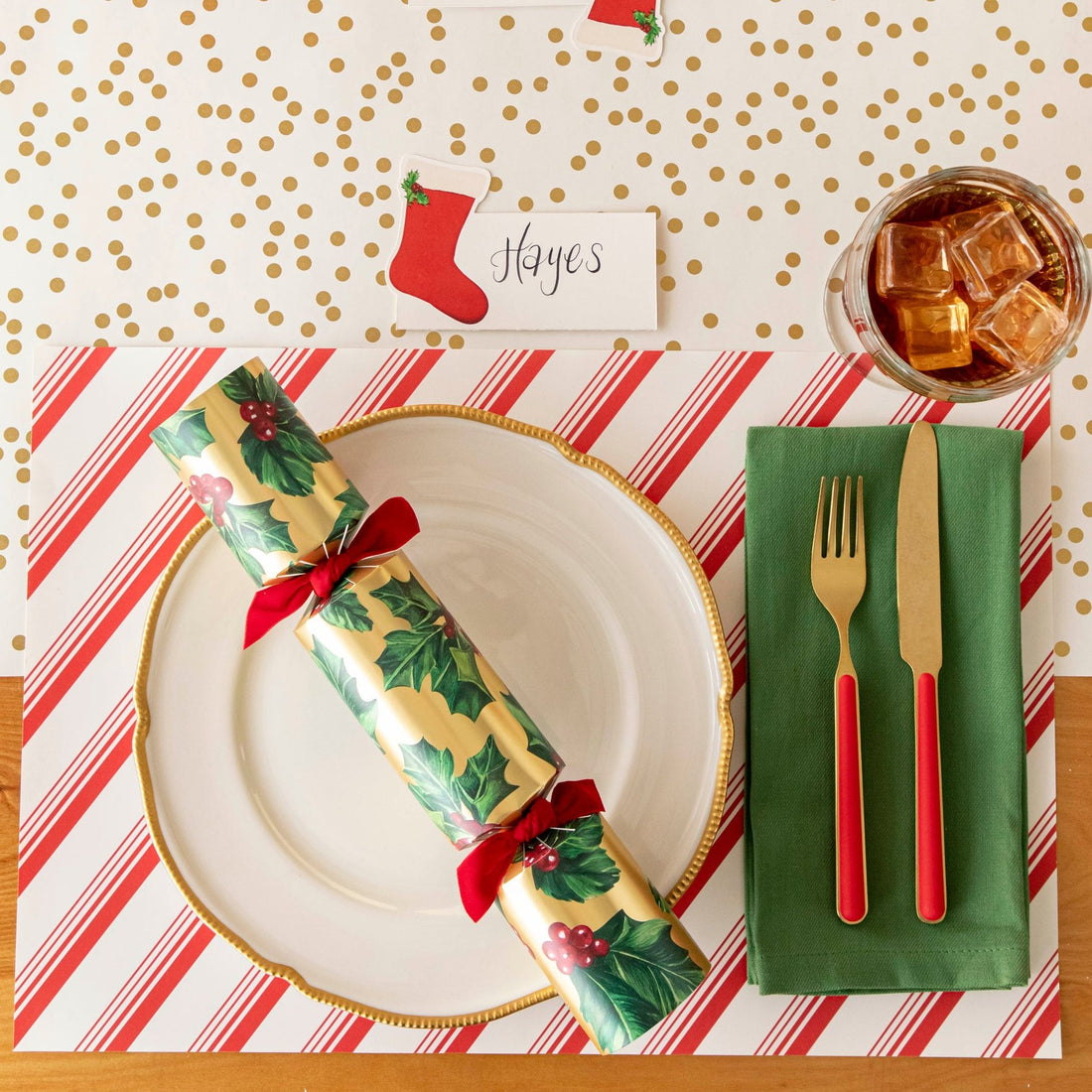 An elegant Christmas themed table setting featuring the Peppermint Stripe Placemat under a gold rimmed dinner plate, green napkin, gold and red silverware, and a Gold Holly Cracker on top.