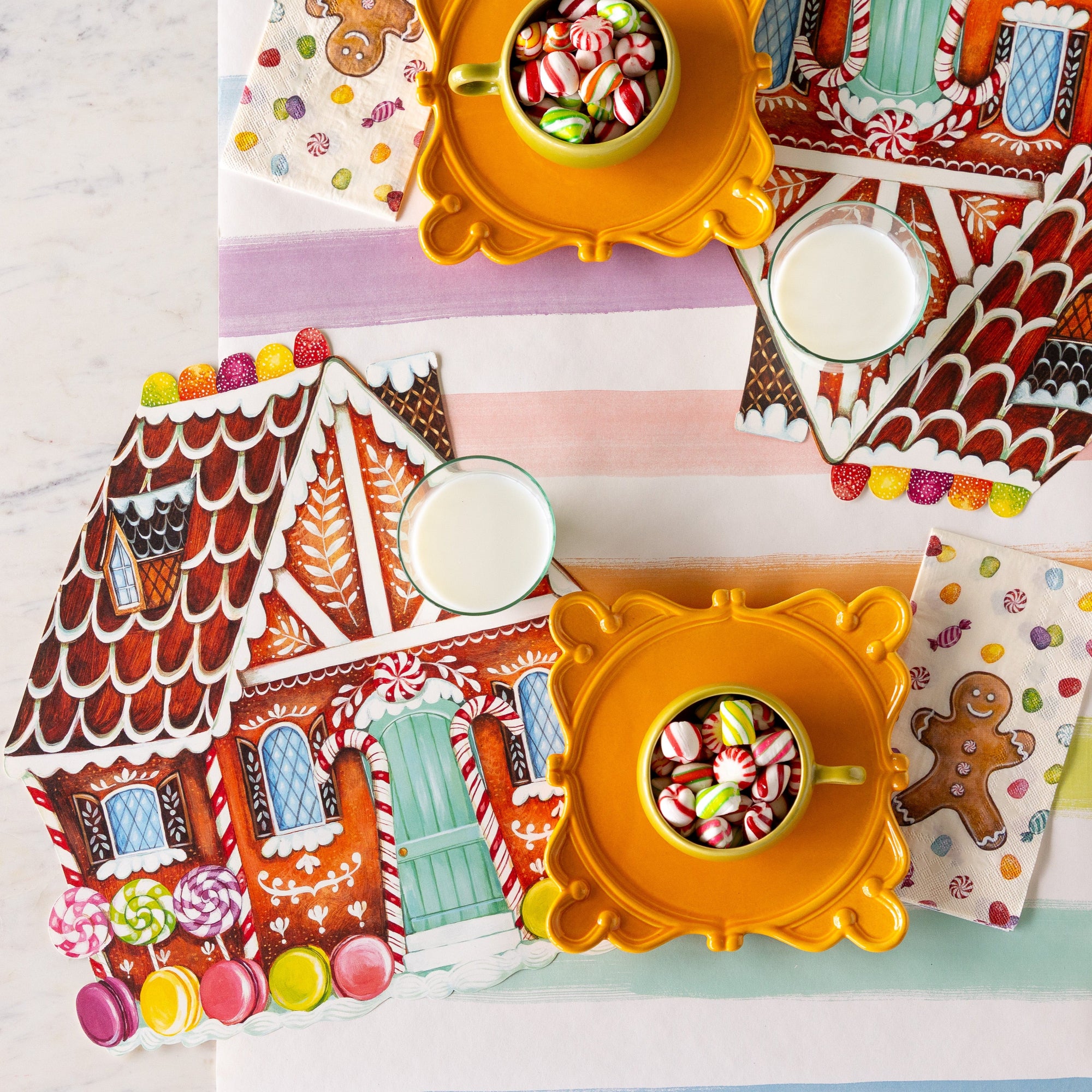 The Die-cut Gingerbread House Placemat with a glass of milk atop, next to a mug of candy on a pedestal and Gingerbread Guest Napkin on the Sorbet Painted Stripe Runner.