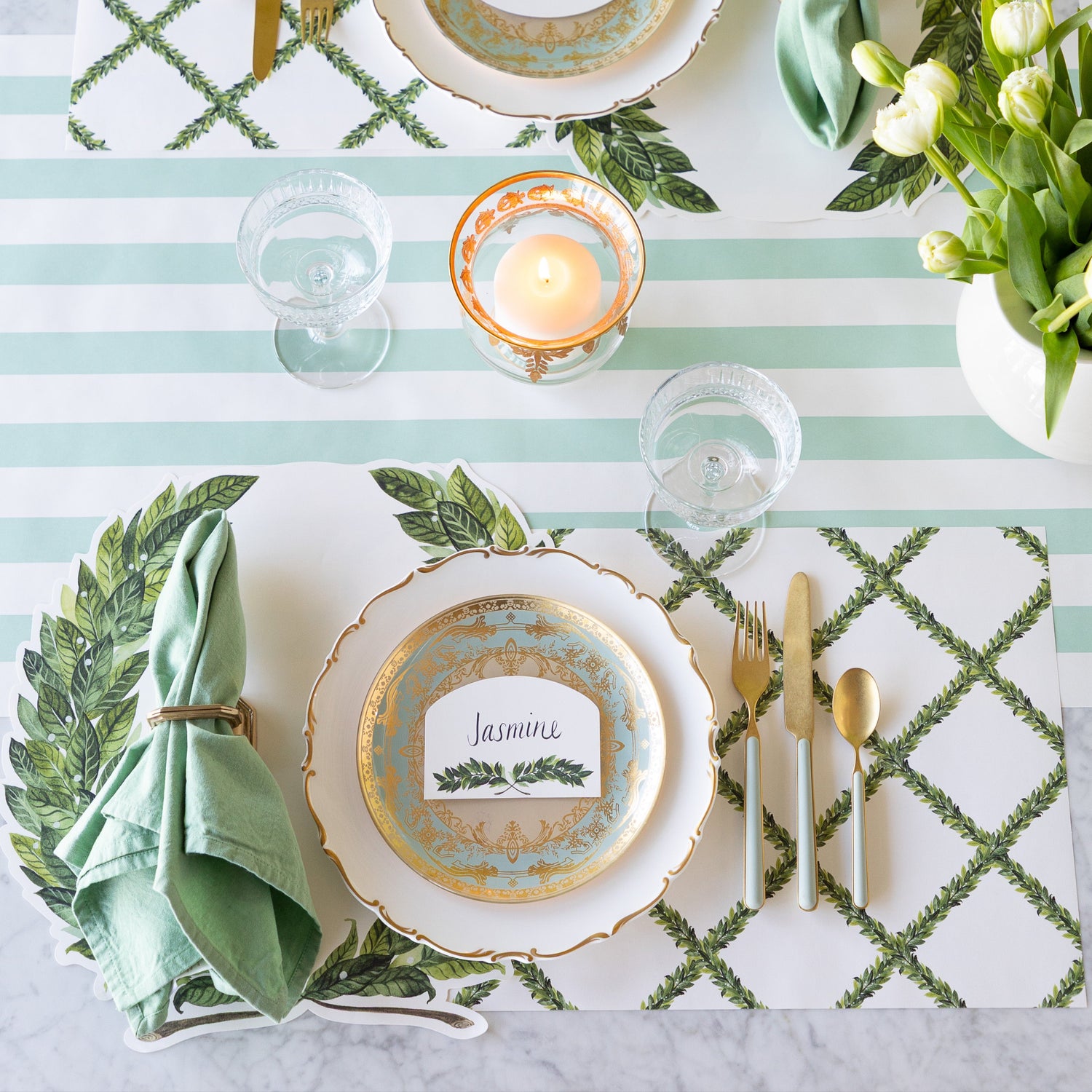 A table setting with a decorative plate, gold utensils, a green napkin in a ring, a glass, and candle sits atop the Die-cut Laurel Placemat and Laurel Lattice Placemat, with the Laurel place card labeled &quot;Jasmine&quot;, on the Seafoam Classic Stripe Runner.