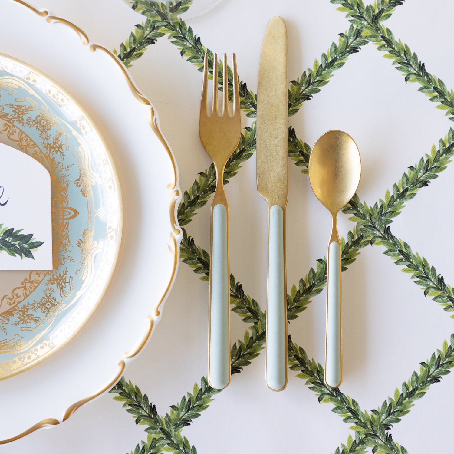 A place setting with decorative plates and gold flatware with seafoam handles, sit atop the Laurel Lattice Placemat.