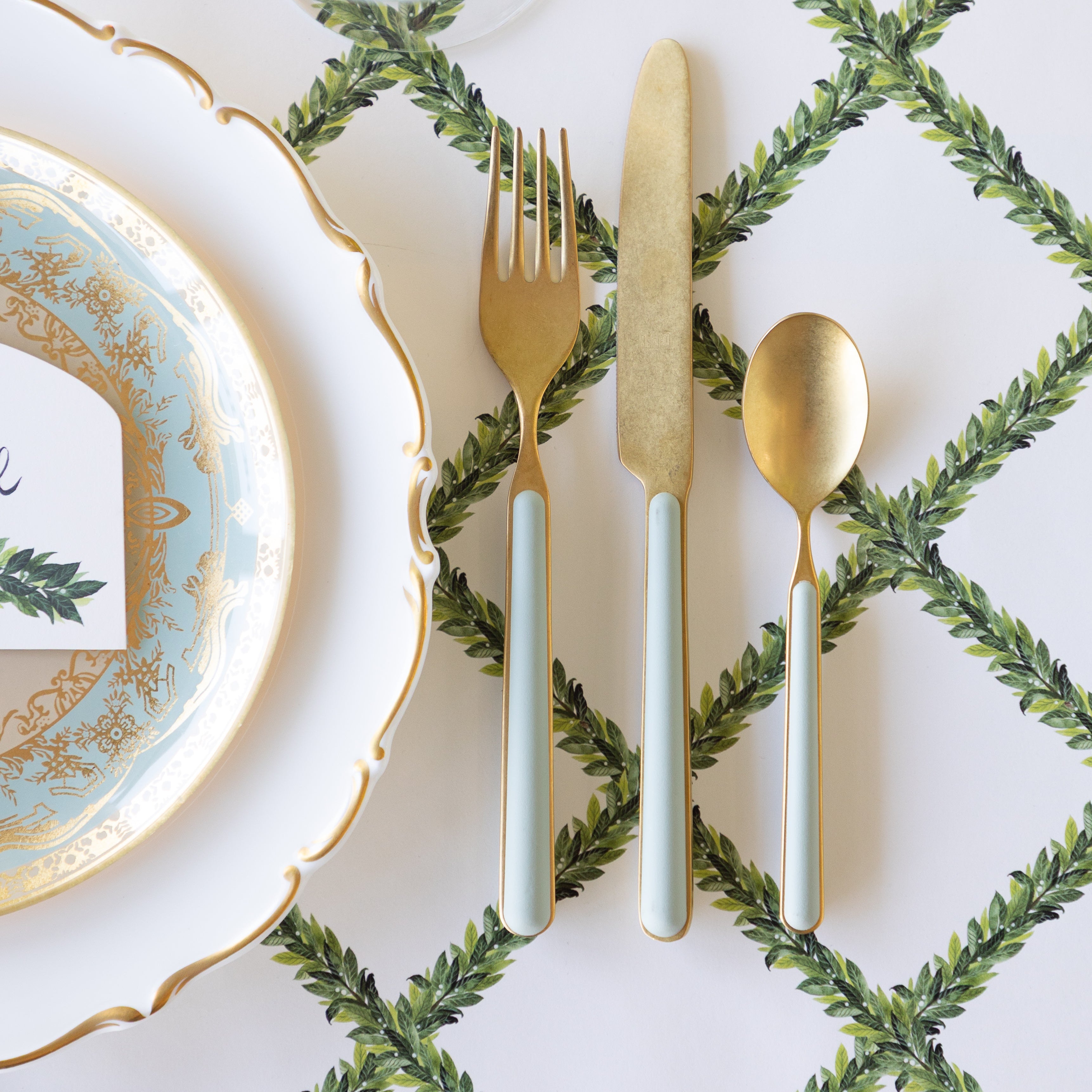 A place setting with decorative plates and gold flatware with seafoam handles, sit atop the Laurel Lattice Placemat.