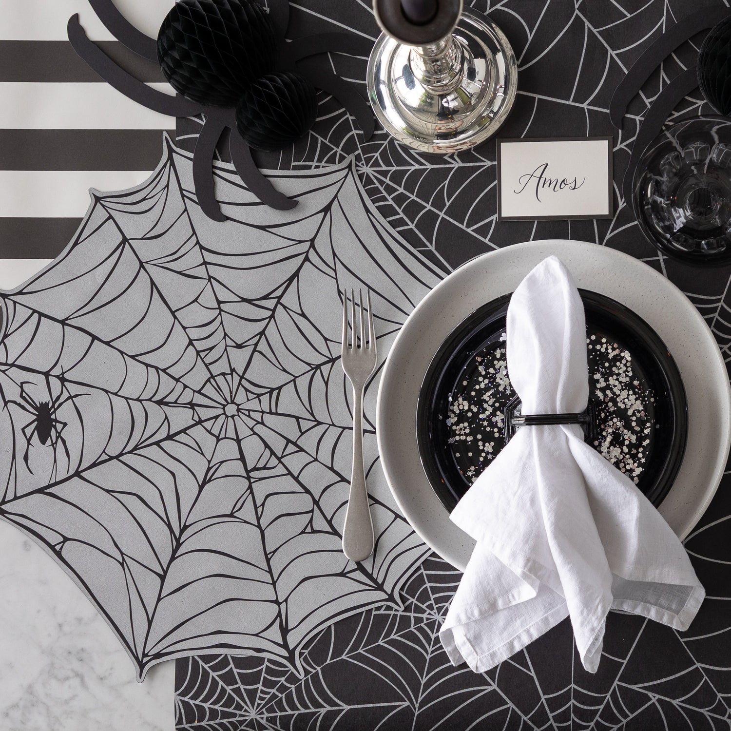 A Halloween-themed place setting with the Die-cut Spiderweb Placemat, a honeycomb spider and Black Frame Place Card reading "Amos" on the Spiderweb Runner.