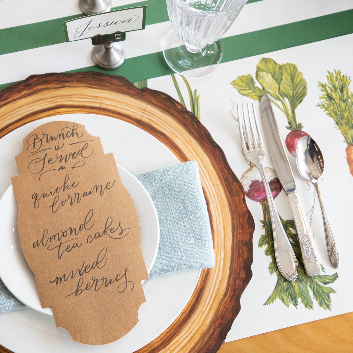 A Die-cut Wood Slice placemat on top of a Dark green classic stripe runner and a vegetable themed placemat underneath with white dinner plates, a seafoam napkin and a Kraft Table Accent on top.