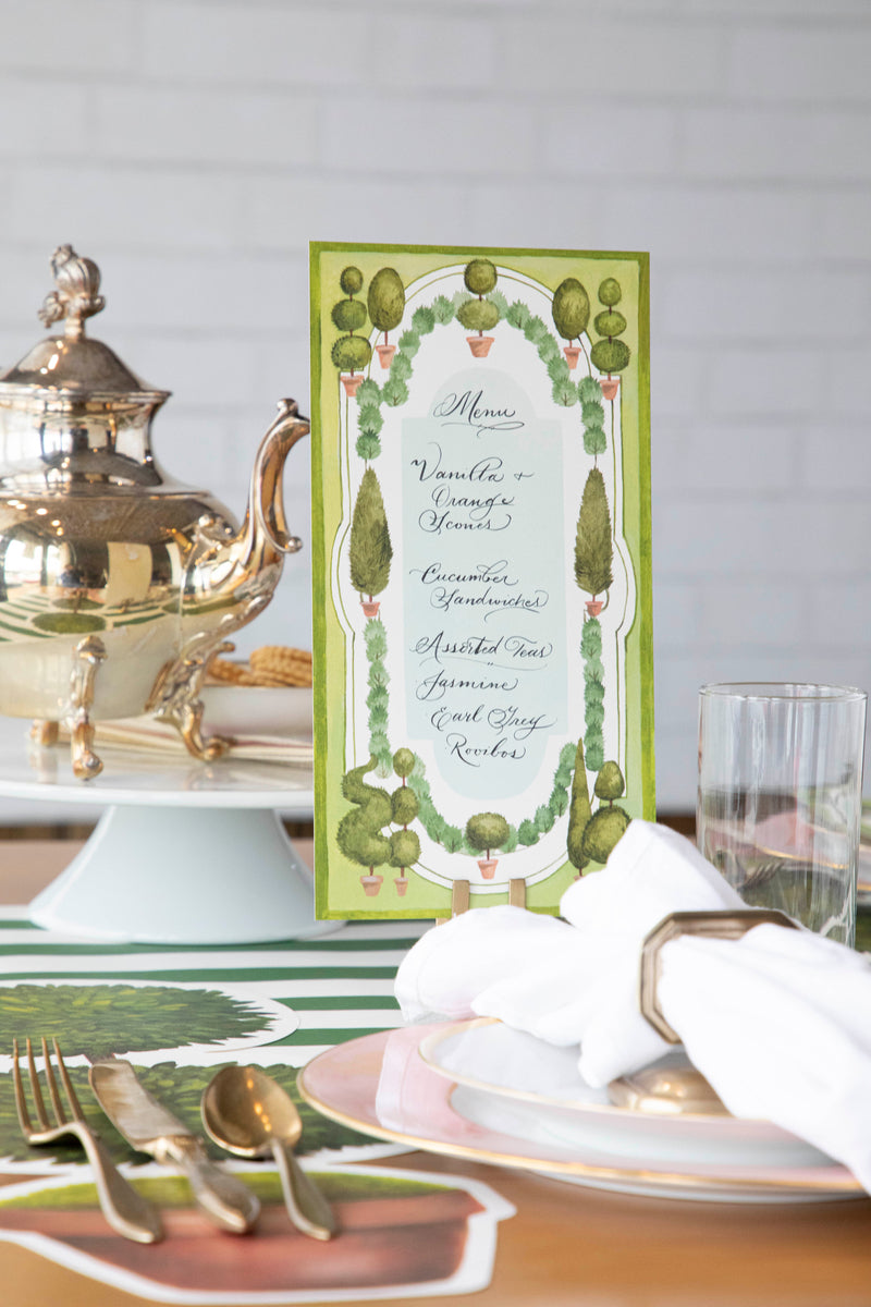 An elegant place setting featuring a Topiary Garden Table Card with a menu written on it standing in a place card holder.