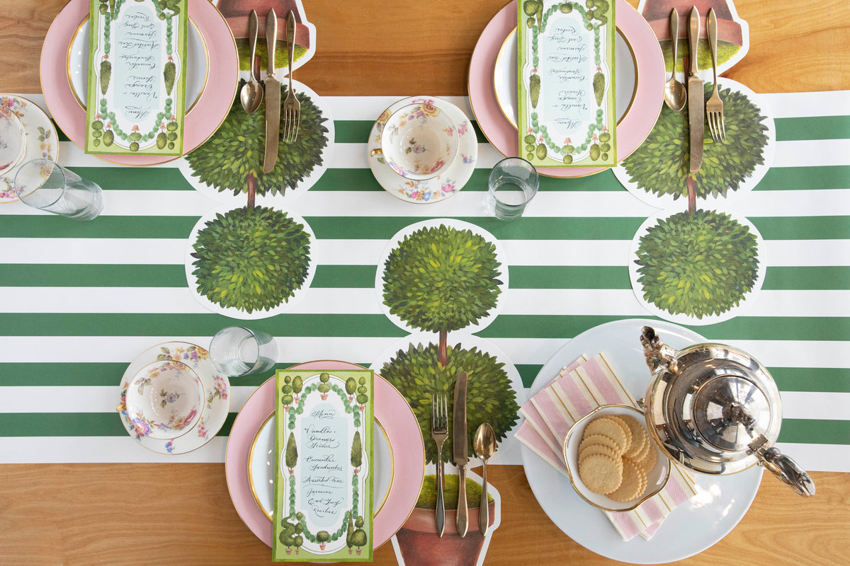A table setting with a green and white striped tablecloth, accompanied by Pink &amp; Gold Awning Stripe Napkins from Hester &amp; Cook for a festive party atmosphere.
