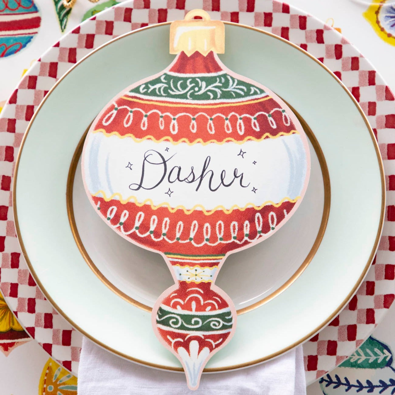 A red Ornaments Table Accent labeled &quot;Dasher&quot; resting on the plate of a Christmas-themed place setting.