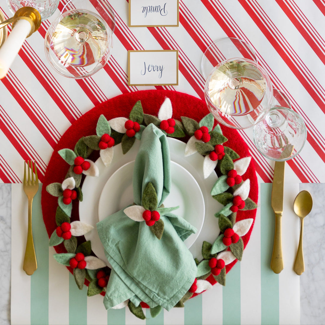 A place setting featuring the Red Felt Placemat, a mint napkin tied in the Felt Berry Napkin Ring and Felt Berry Charger atop, Gold Frame Place Card, and gold flatware on the Candy Stripe Runner and Seafoam Classic Stripe Runner.