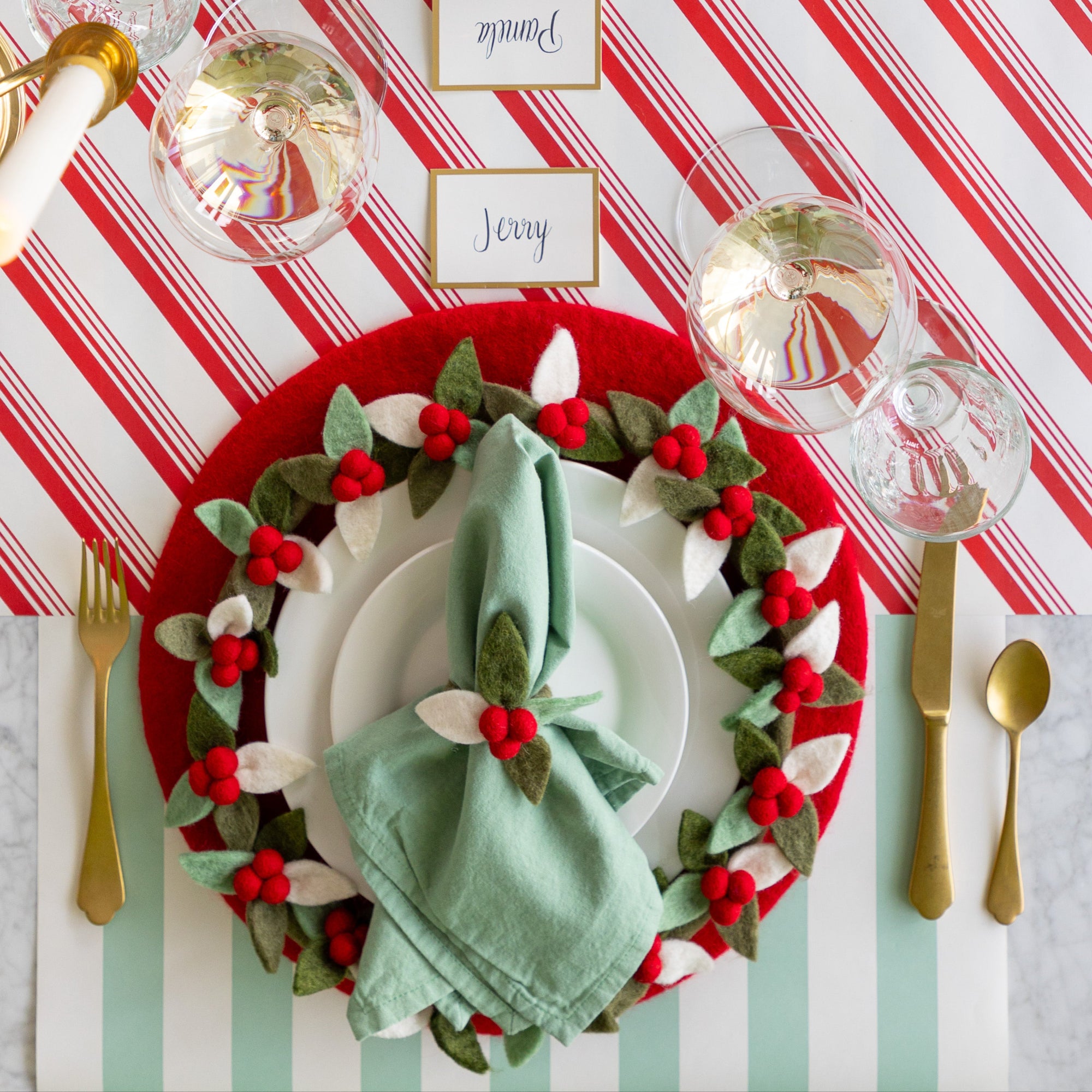 A place setting featuring the Red Felt Placemat, a mint napkin tied in the Felt Berry Napkin Ring and Felt Berry Charger atop, Gold Frame Place Card, and gold flatware on the Candy Stripe Runner and Seafoam Classic Stripe Runner.