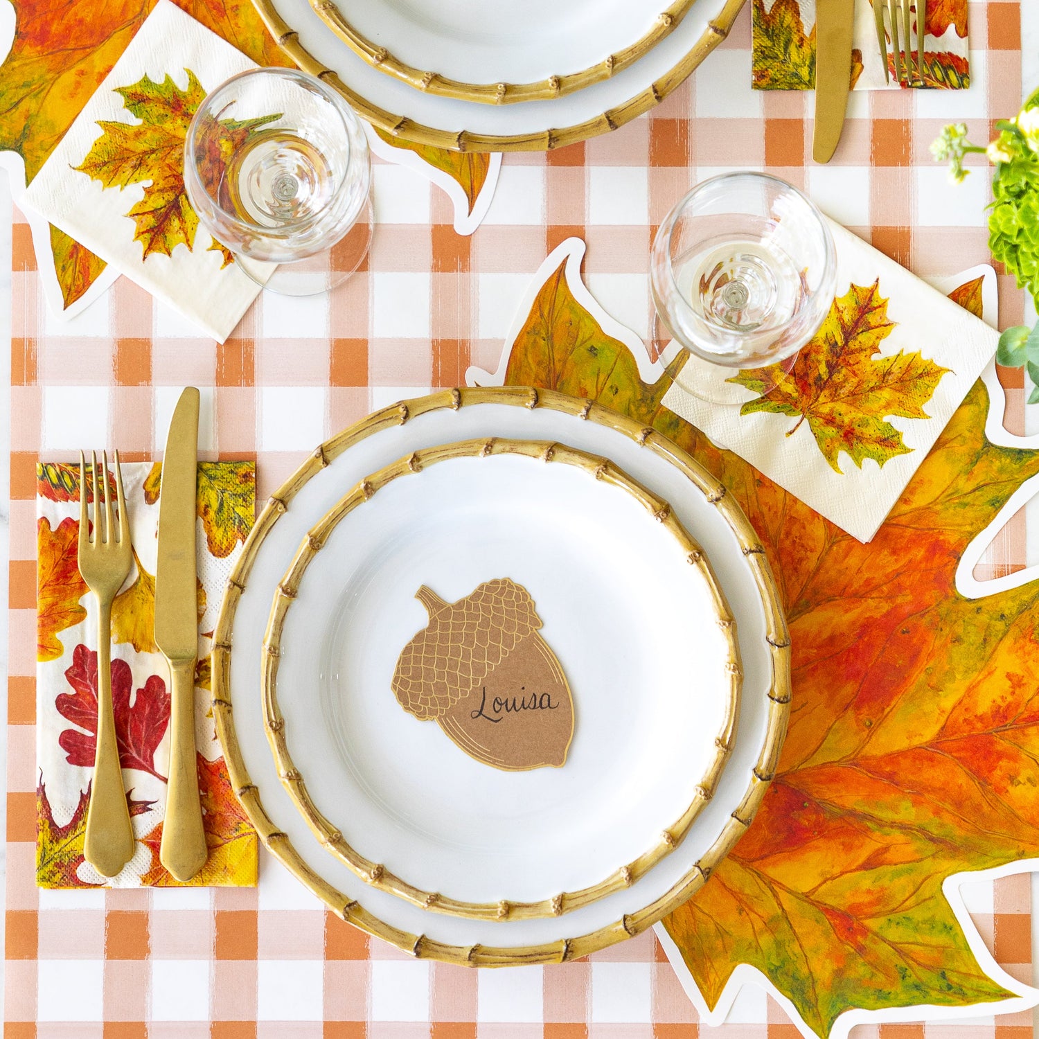 An elegant place setting featuring the Die-cut Fall Leaf Placemat, Juliska Bamboo plates, the Acorn Place Card reading &quot;Louisa&quot;, Fall Foliage Cocktail and Guest Napkins and the Orange Painted Check Runner underneath.