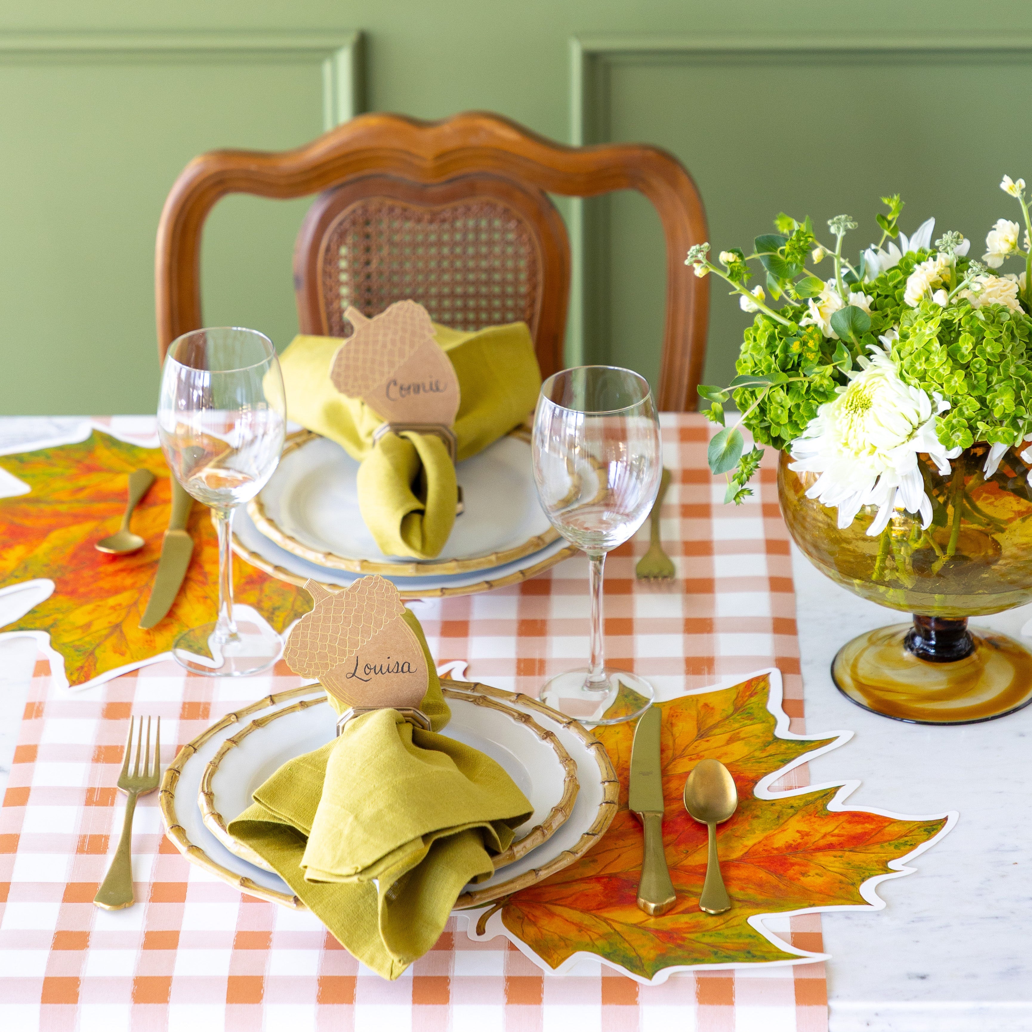 A elegant table setting with flowers, featuring the Die-cut Fall Leaf Placemat with the Napkin Ring with Place Card Holder holding the Acorn Place Card on top of the plates and Orange Painted Check Runner underneath.