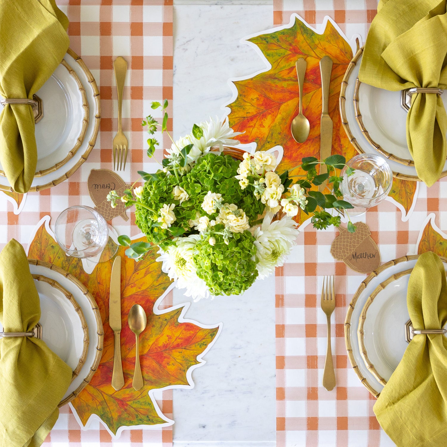 An elegant table setting with flowers in the center, featuring the Die-cut Fall Leaf Placemat under Juliska Bamboo plates, Mepra gold flatware and Acorn Place Card with the Orange Painted Check Runner underneath.