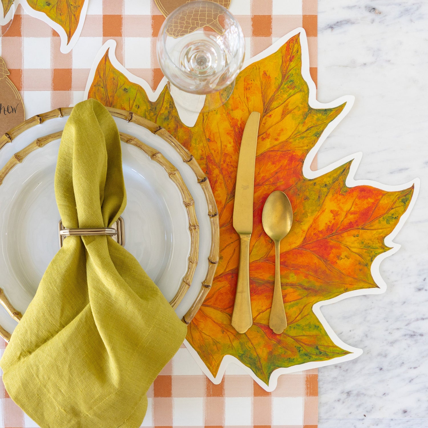 An autumn place setting featuring the Die-cut Fall Leaf Placemat under Juliska Bamboo Dinner Plates with a green napkin in a holder on top, gold flatware and Orange Painted Check Runner underneath.