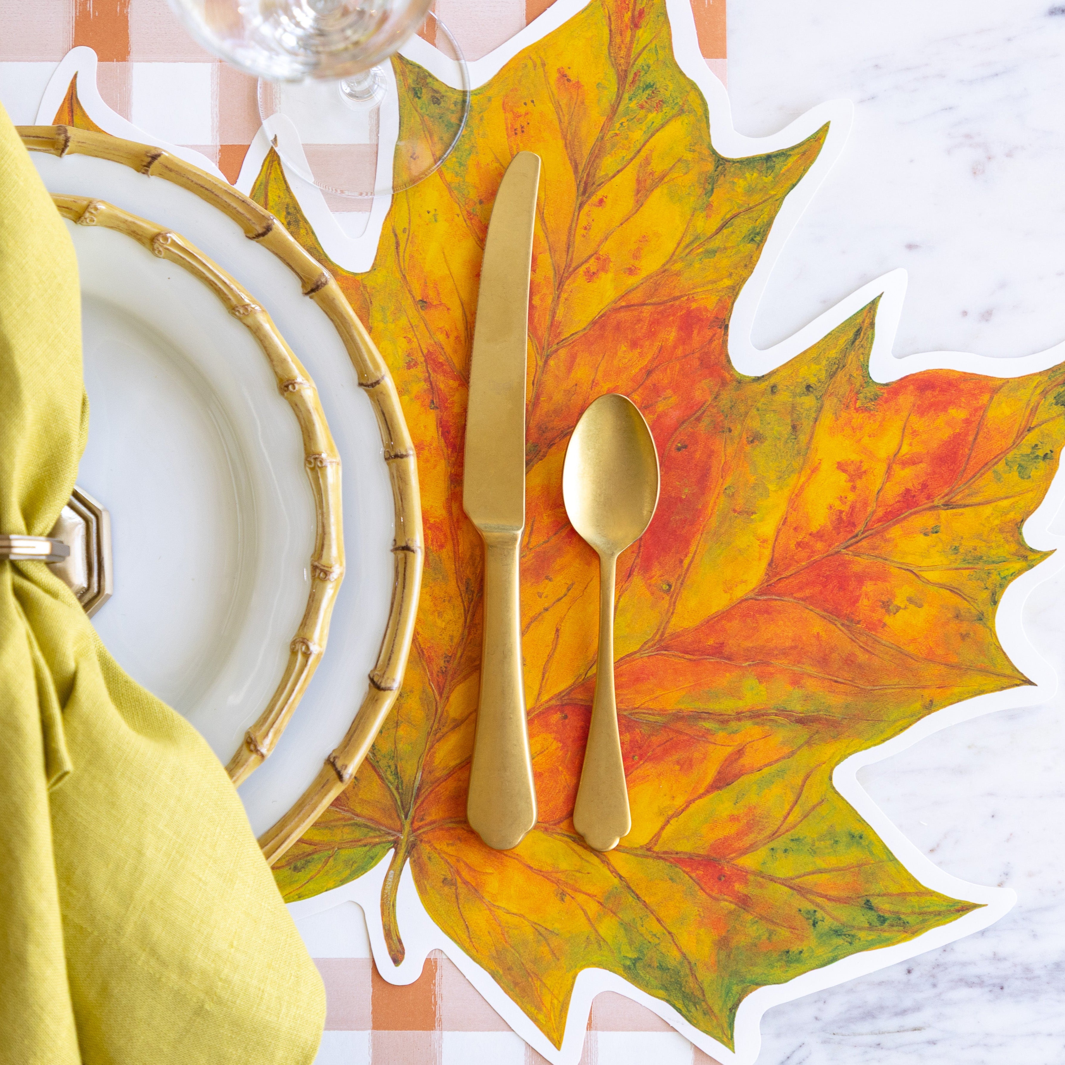 A place setting featuring the Die-cut Fall Leaf Placemat under Juliska Bamboo Dinner plates, a green napkin in a holder, the Mepra gold knife and spoon and Orange Painted Check Runner underneath.