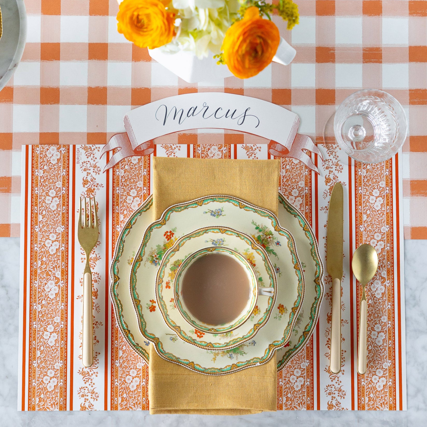A fall place setting featuring the Pumpkin Burleigh Stripe Placemat, vintage floral dinnerware, a tan napkin, Orange Classic Banner Table Accent reading &quot;Marcus&quot;, and gold flatware on the Orange Painted Check Runner.