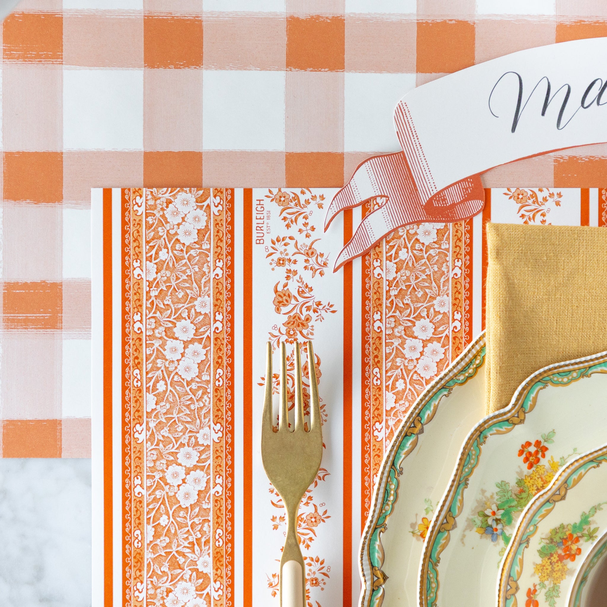 A close up of the Pumpkin Burleigh Stripe Placemat under a place setting, showing the orange stripes and floral design, on the Orange Painted Check Runner.