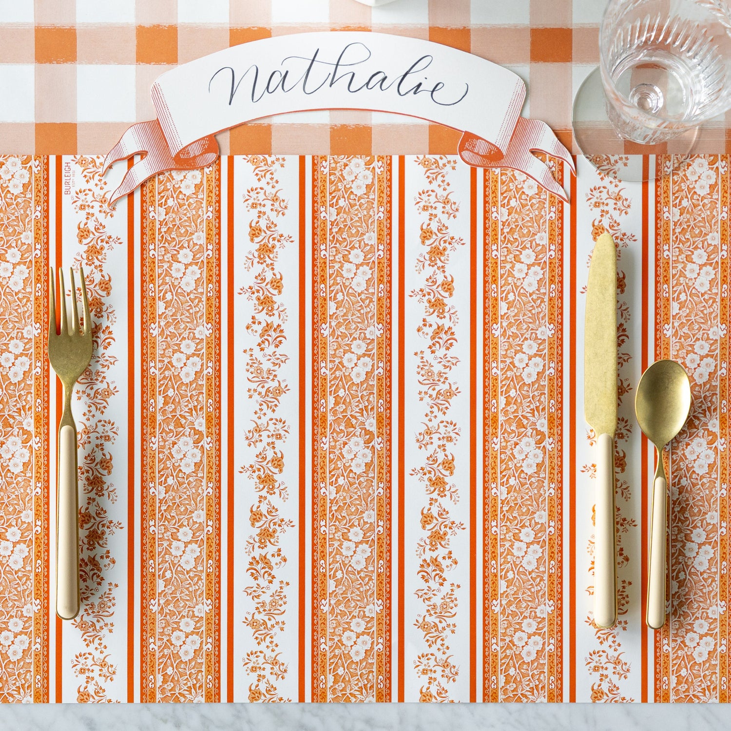 A place setting with the Pumpkin Burleigh Stripe Placemat with gold and peach flatware and Classic Orange Banner Table Accent reading &quot;Nathalie&quot; atop, on the Orange Painted Check Runner.
