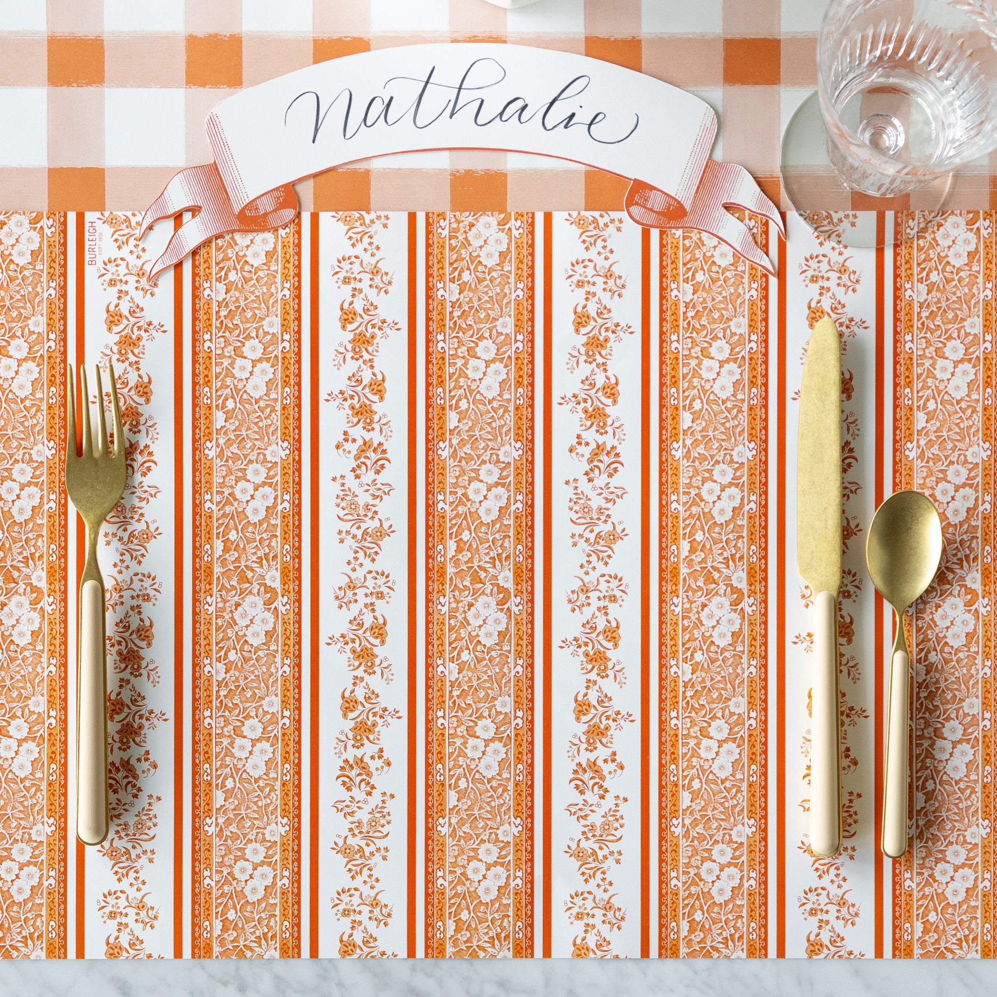 A place setting with the Pumpkin Burleigh Stripe Placemat with gold and peach flatware and Classic Orange Banner Table Accent reading &quot;Nathalie&quot; atop, on the Orange Painted Check Runner.