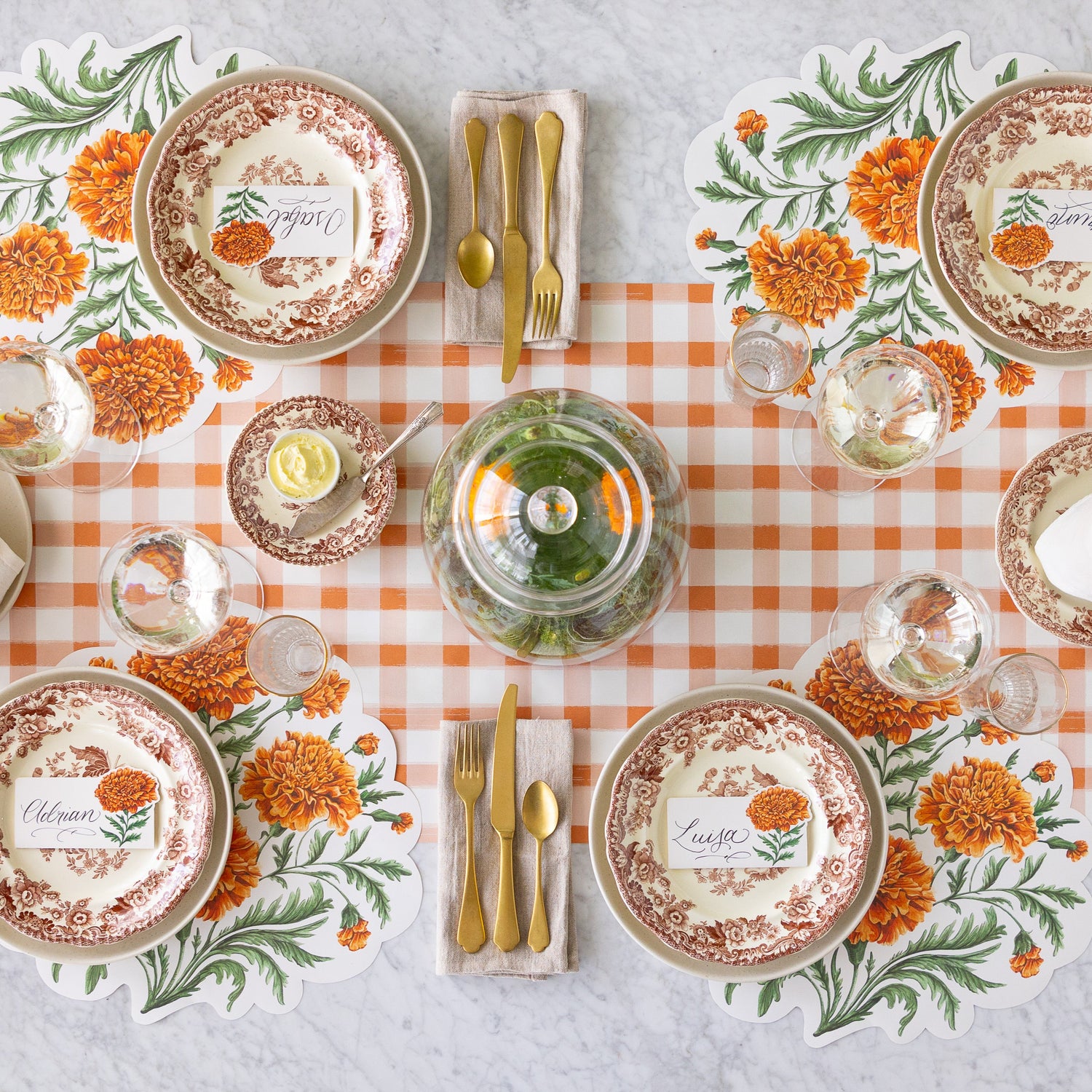 An elegant table setting for four with a cloche of Marigolds in the center, featuring the Die-cut Marigold Bouquet Placemats, vintage dinnerware with the Marigold Bloom Place Cards on top, a wine and water glass and the Orange Painted Check Runner underneath.