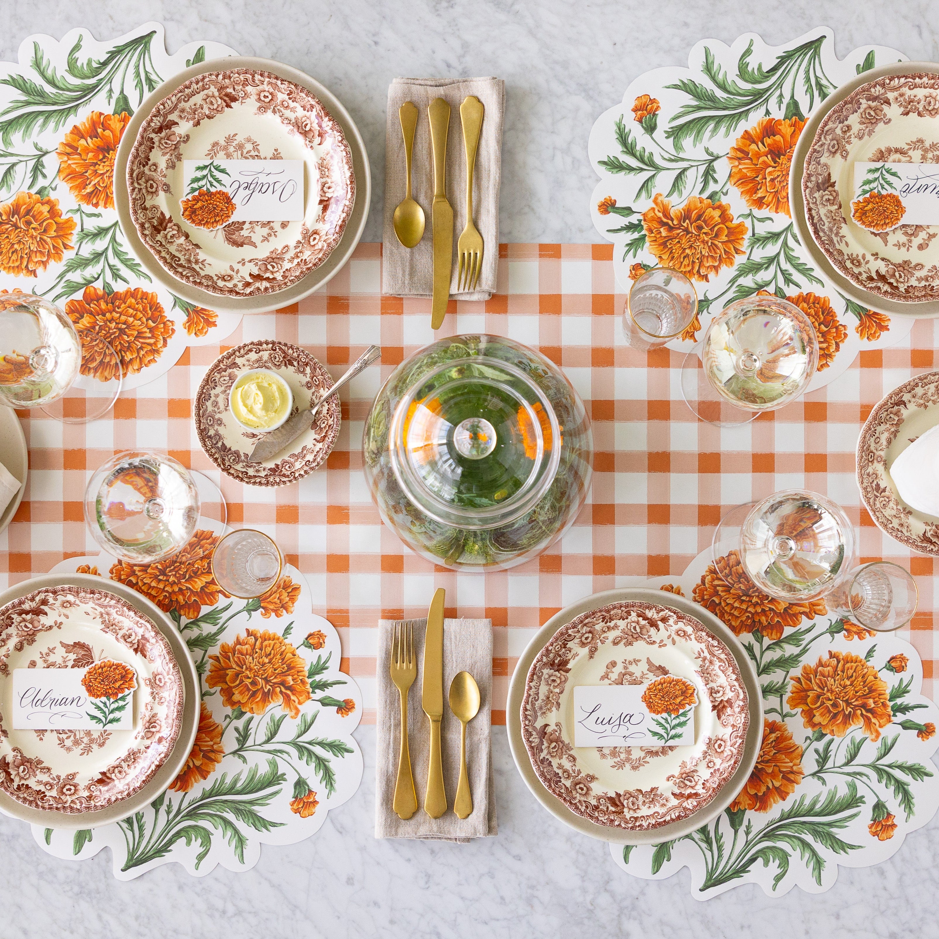 An elegant table setting for four with a cloche of Marigolds in the center, featuring the Die-cut Marigold Bouquet Placemats, vintage dinnerware with the Marigold Bloom Place Cards on top, a wine and water glass and the Orange Painted Check Runner underneath.