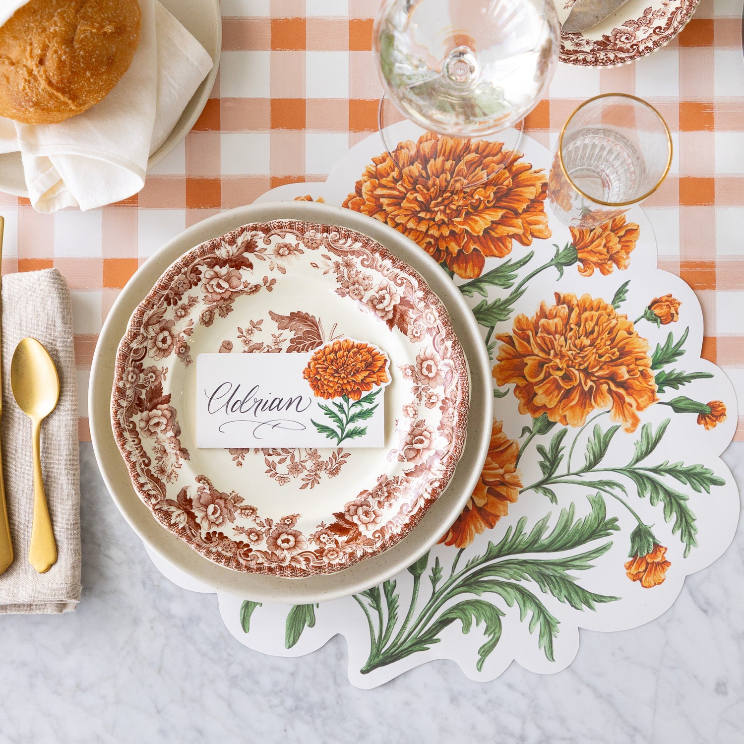 An elegant table setting featuring the Die-cut Marigold Bouquet Placemat under vintage floral dinnerware with the Marigold Bloom Place Card on top, a wine and water glass, Mepra Gold Flatware on a napkin nearby, and the Orange Painted Check Runner underneath.