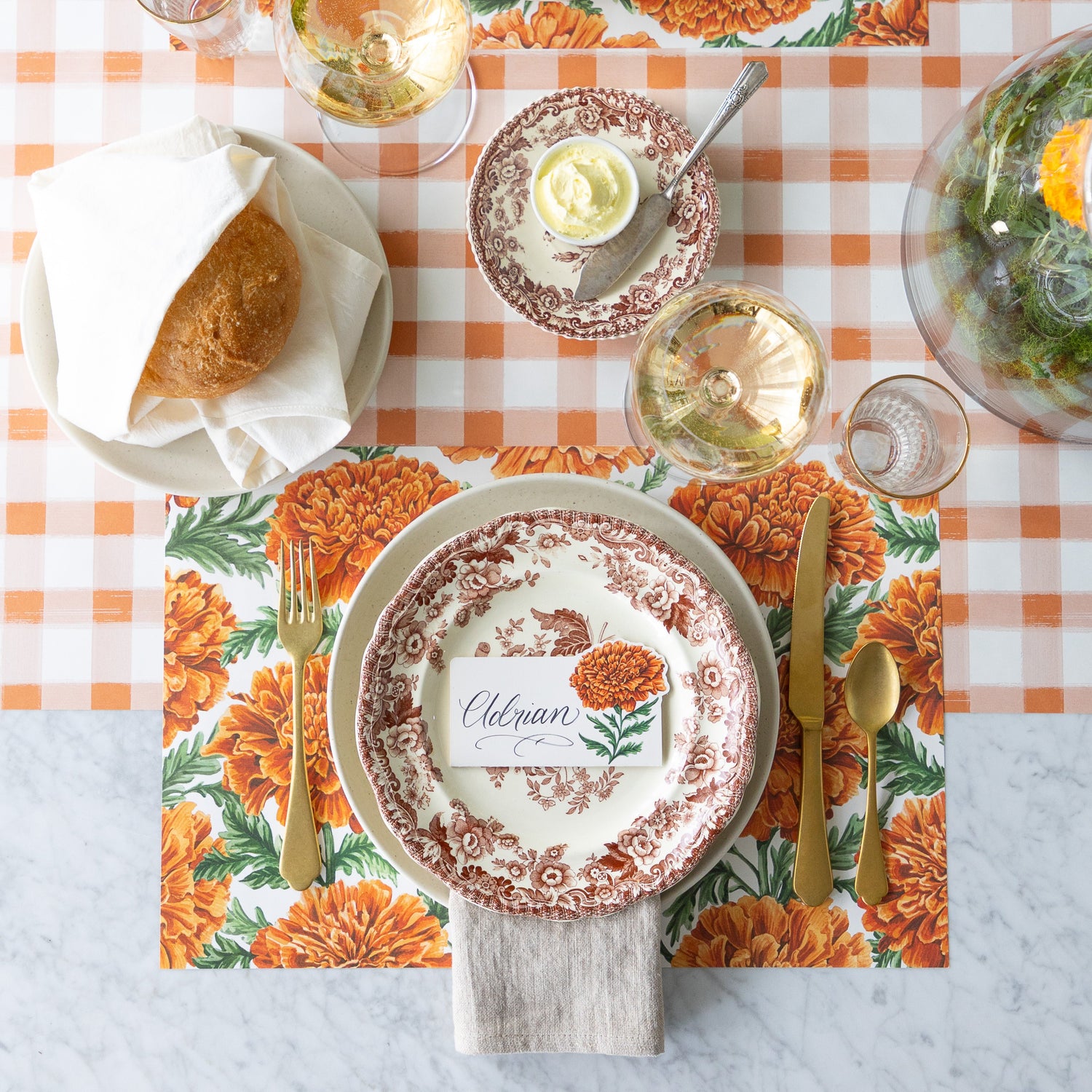 An elegant table setting with a vintage floral plate, gold utensils, bread roll, wine glass, and a Marigold Bloom Place Card reading &quot;Eleanor&quot; atop a Marigold Harvest Placemat—on a Orange Painted Check Runner.