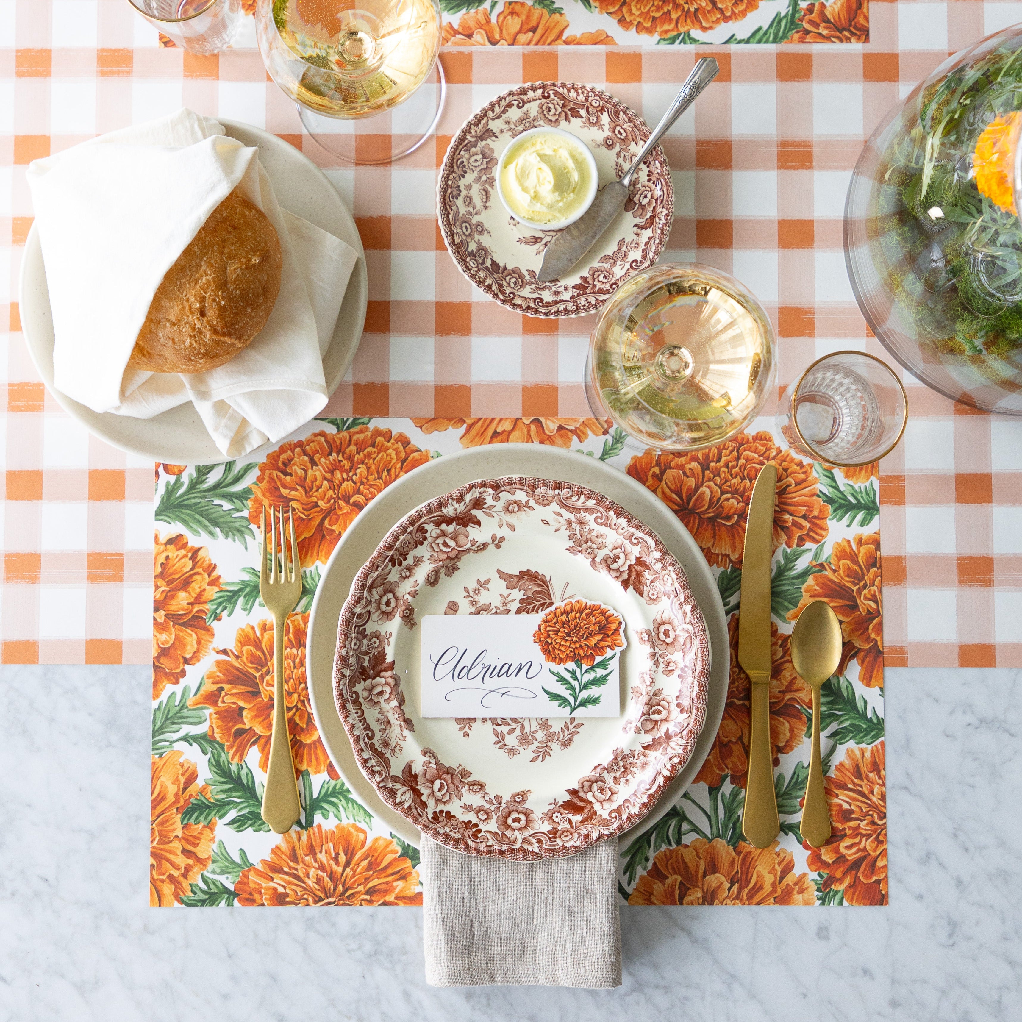 An elegant table setting with a vintage floral plate, gold utensils, bread roll, wine glass, and a Marigold Bloom Place Card reading &quot;Eleanor&quot; atop a Marigold Harvest Placemat—on a Orange Painted Check Runner.