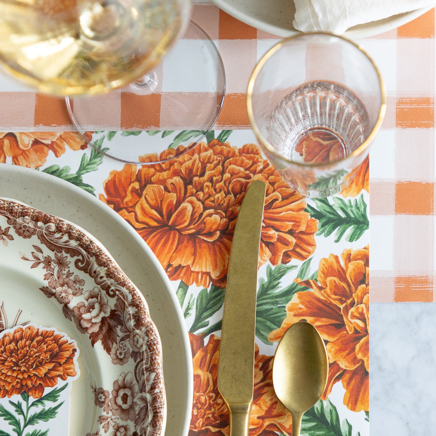 An elegant place setting featuring the Marigold Harvest Placemat under dinnerware, gold flatware, and a water and wine glass, with the Orange Painted Check Runner underneath.