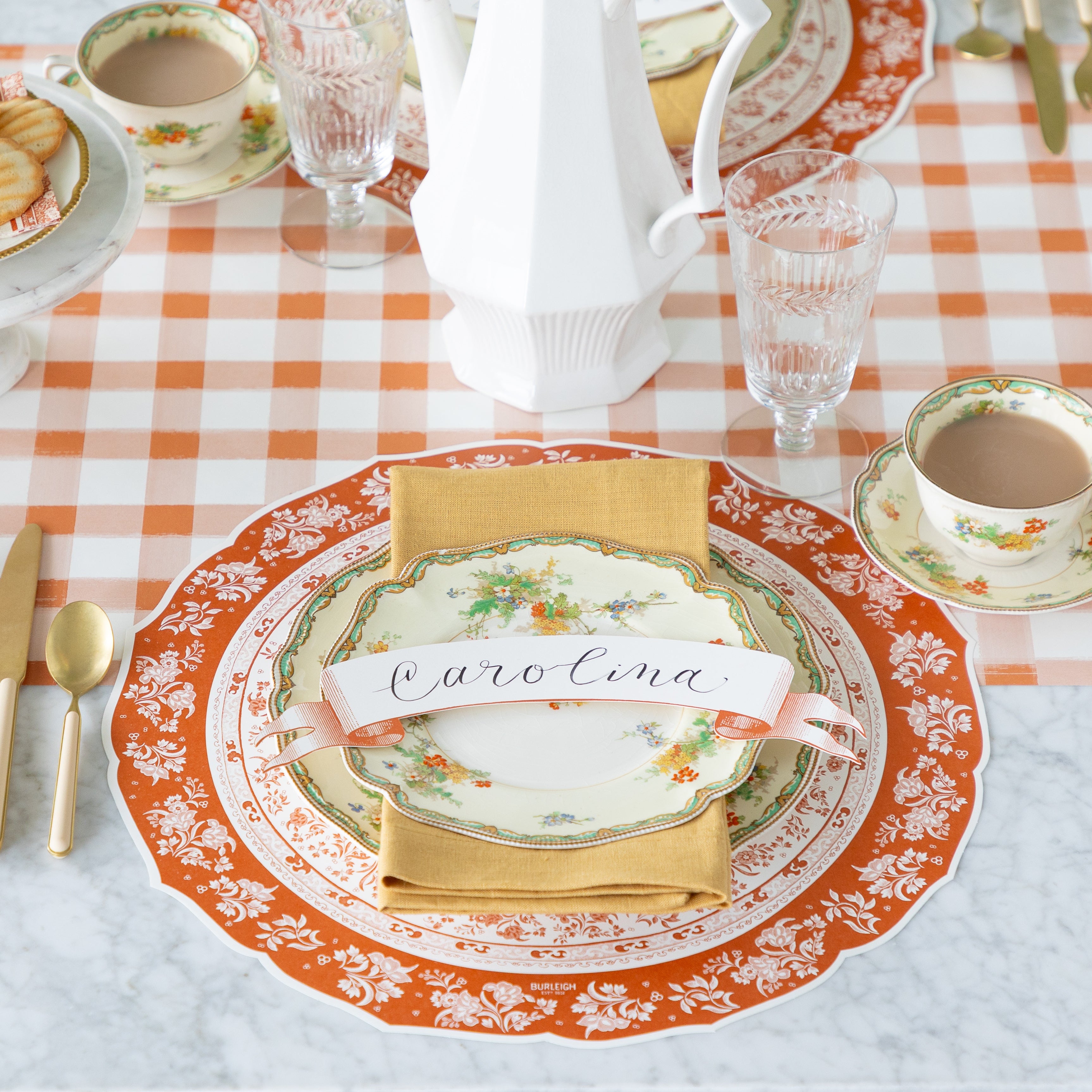 A fall place setting featuring the Die-cut Pumpkin Regal Placemat, vintage floral dinnerware with a tan napkin and Orange Banner Table Accent reading &quot;Carolina&quot;, atop, and a cup of coffee on the Orange Painted Check Runner.
