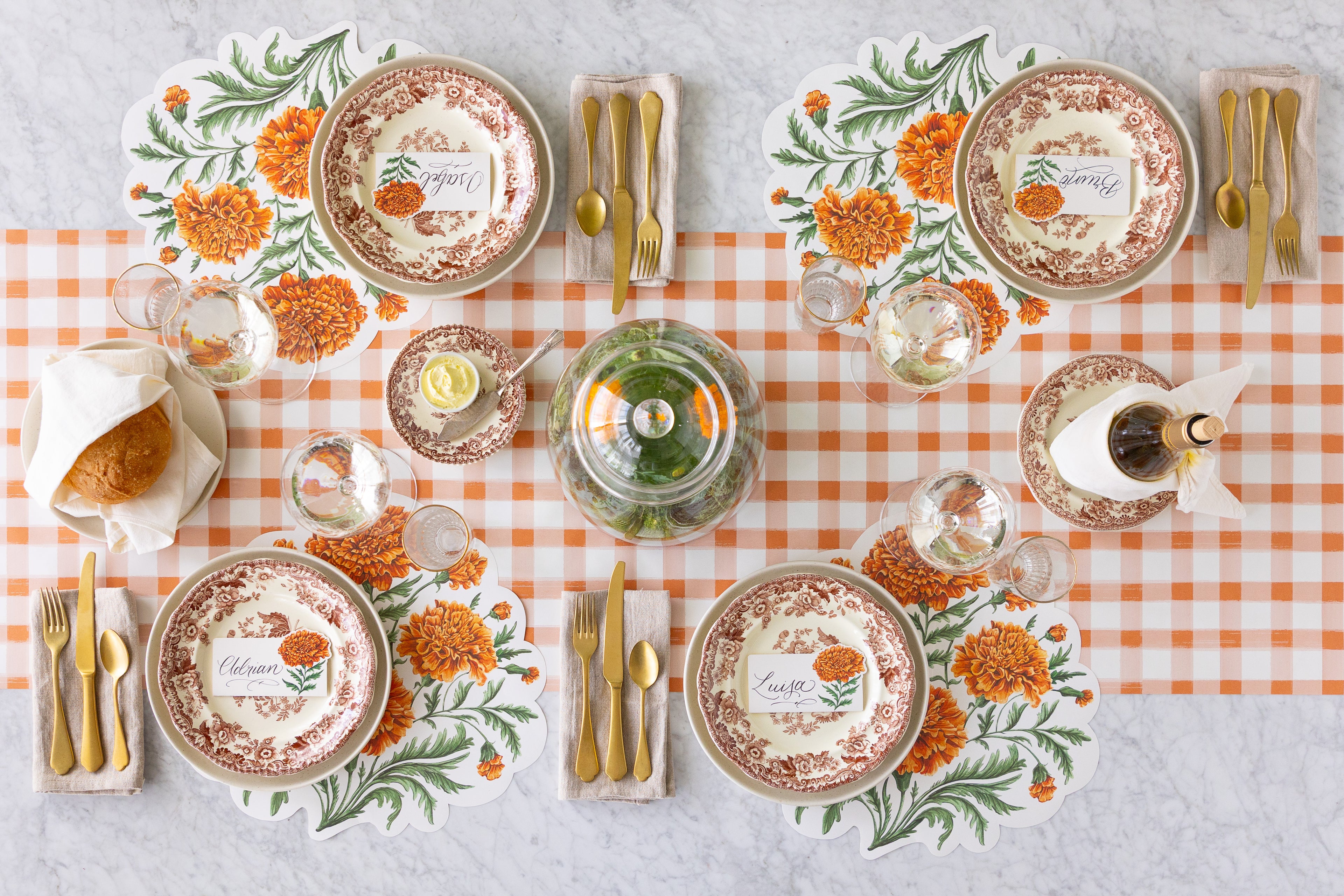 An autumnal table setting featuring the Die-cut Marigold Placemats, floral plates, Marigold Place Cards, gold flatware, glasses of wine and water with a Marigold centerpiece on the Orange Painted Check Runner.