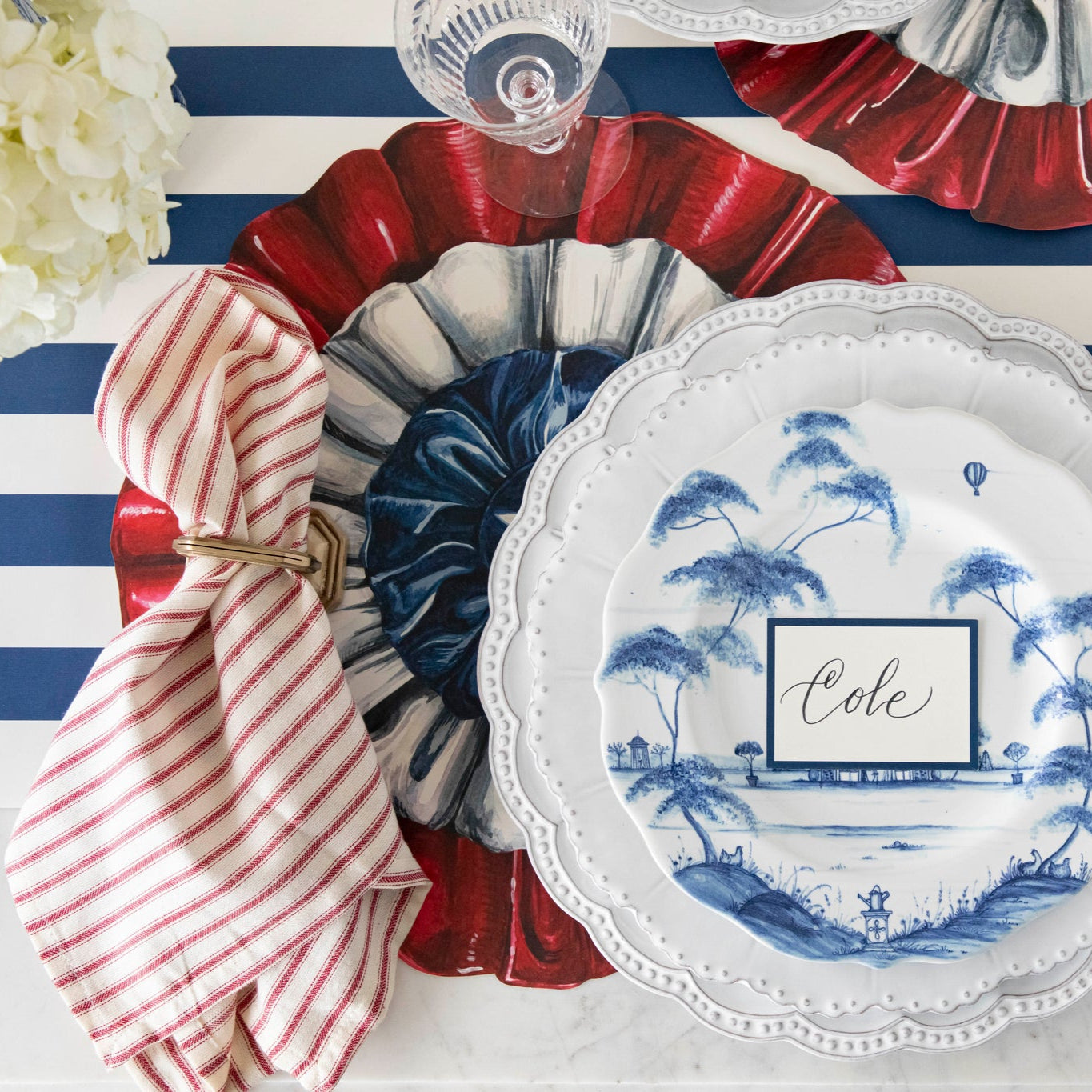 Patriotic table setting featuring the Die-cut Star Spangled Placemat, a red-and-white striped napkin, beaded and blue floral plates, and Blue Frame Place Card on the Navy Classic Stripe Runner.