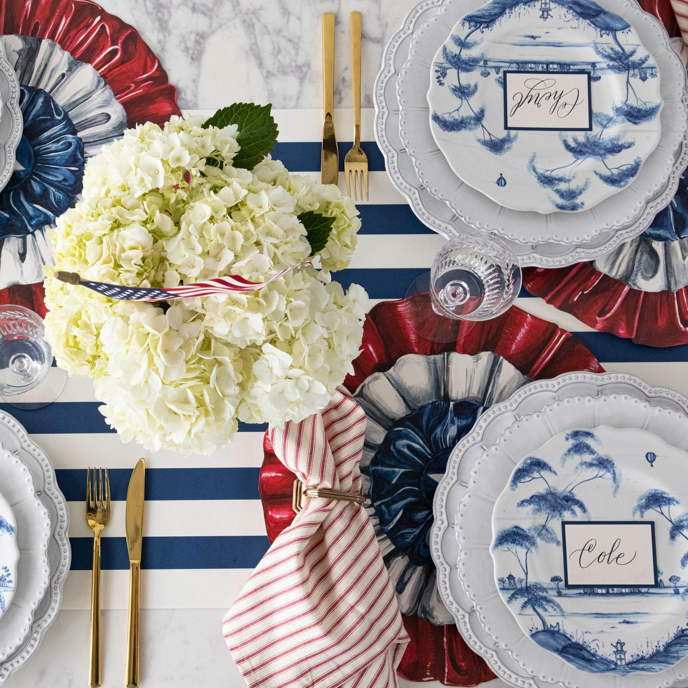 Patriotic table setting featuring the Die-cut Star Spangled Placemat, a red-and-white striped napkin, beaded and blue floral plates, and Blue Frame Place Card on the Navy Classic Stripe Runner.