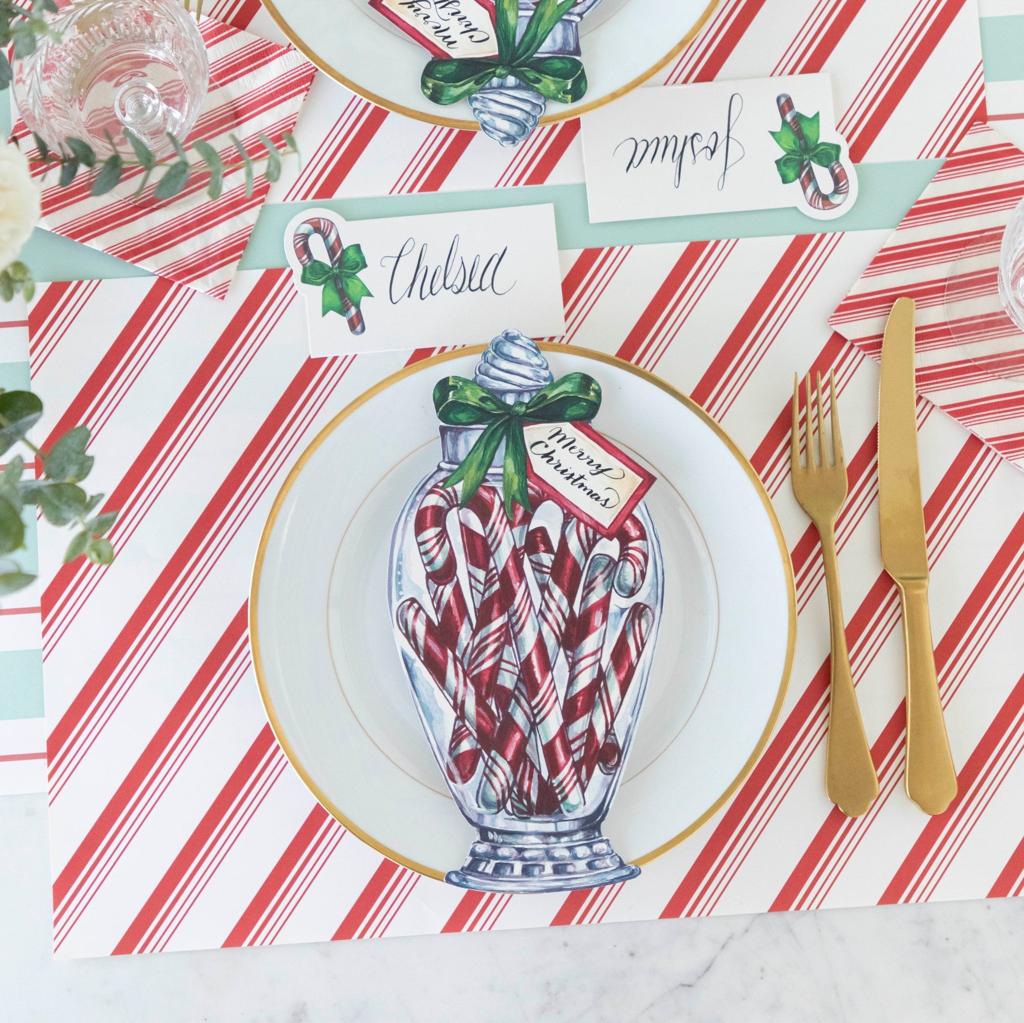 An elegant table setting with the Candy Cane Jar Table Accent with &quot;Merry Christmas&quot; written on the tag on a gold rimmed plate that sits on the Peppermint Stripe Placemat.