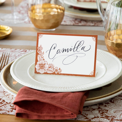 A free-standing, rectangular white table card with a dark orange frame around the edges, embellished with dark orange linework flower designs in the corners.