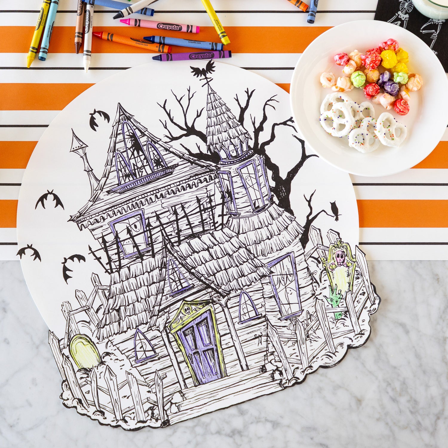 The Die-cut Haunted House Coloring Placemat partially colored in, surrounded by crayons and a bowl of treats, with the Orange &amp; Black Awning Stripe Runner underneath.