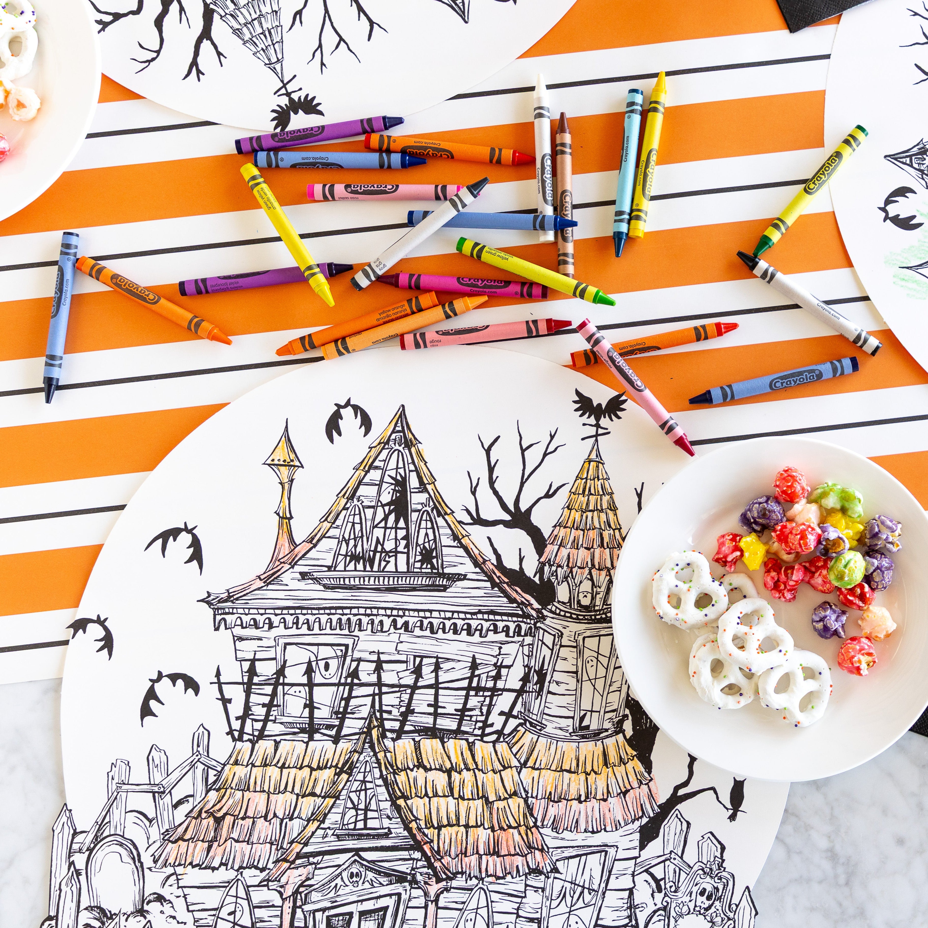 The Die-cut Haunted House Coloring Placemat partially colored in, surrounded by crayons and a bowl of treats, with the Orange &amp; Black Awning Stripe Runner underneath.