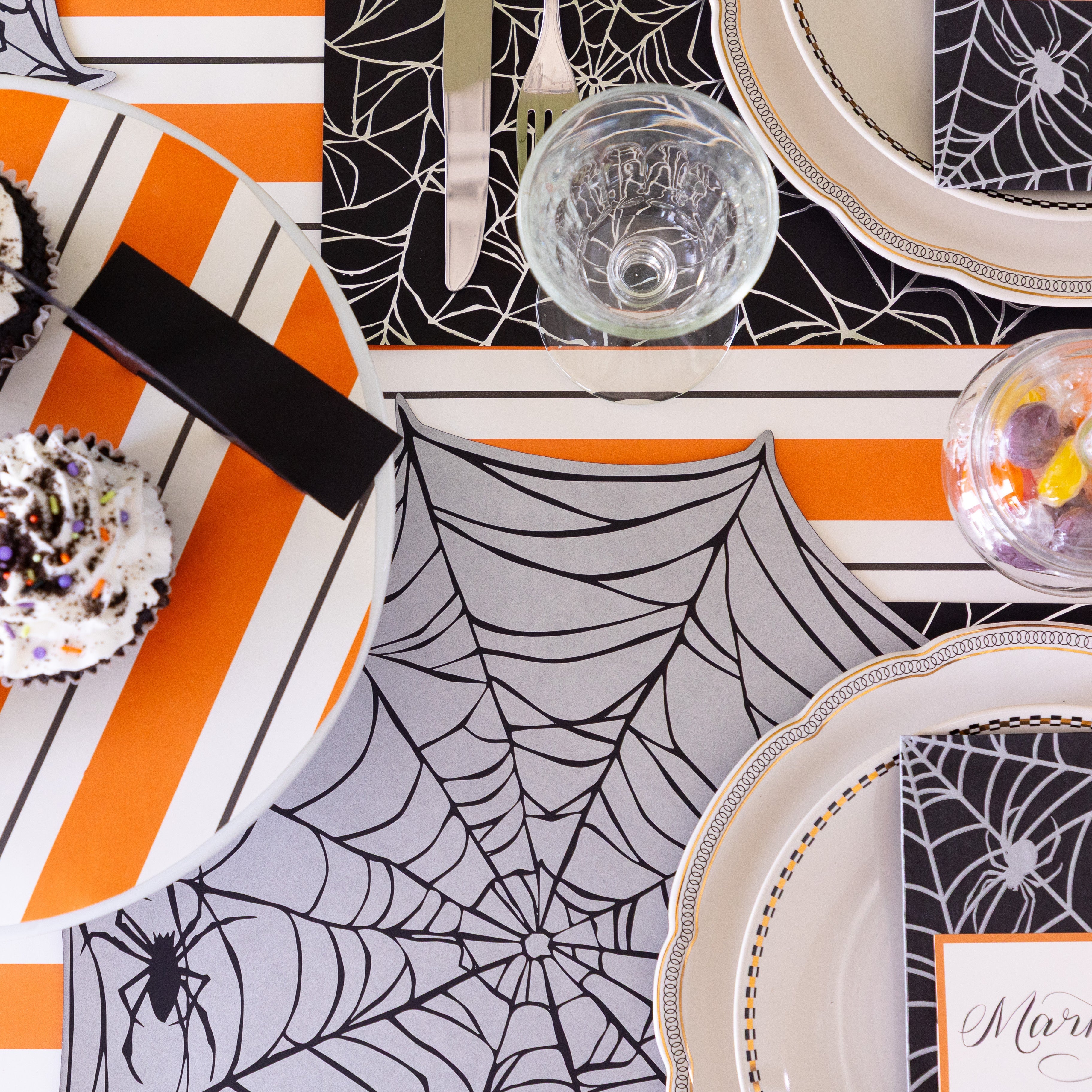 A Halloween themed table setting with halloween themed cupcakes and candy, featuring the Die-cut Spiderweb Placemat on the Orange &amp; Black Awning Stripe Runner.